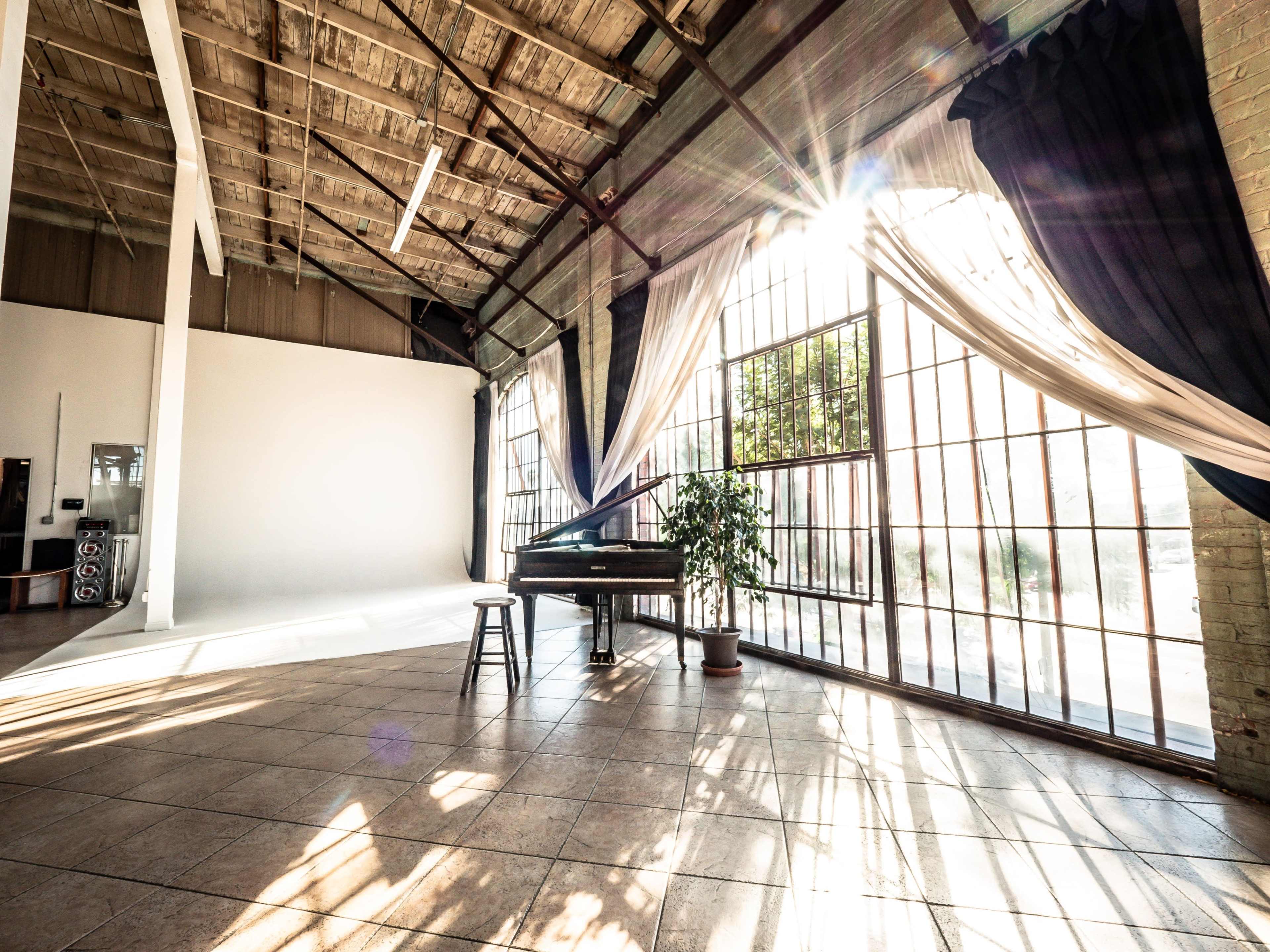 An open, sunlit space featuring large windows, a grand piano, and a stool, accented by draped fabric and a potted plant.
