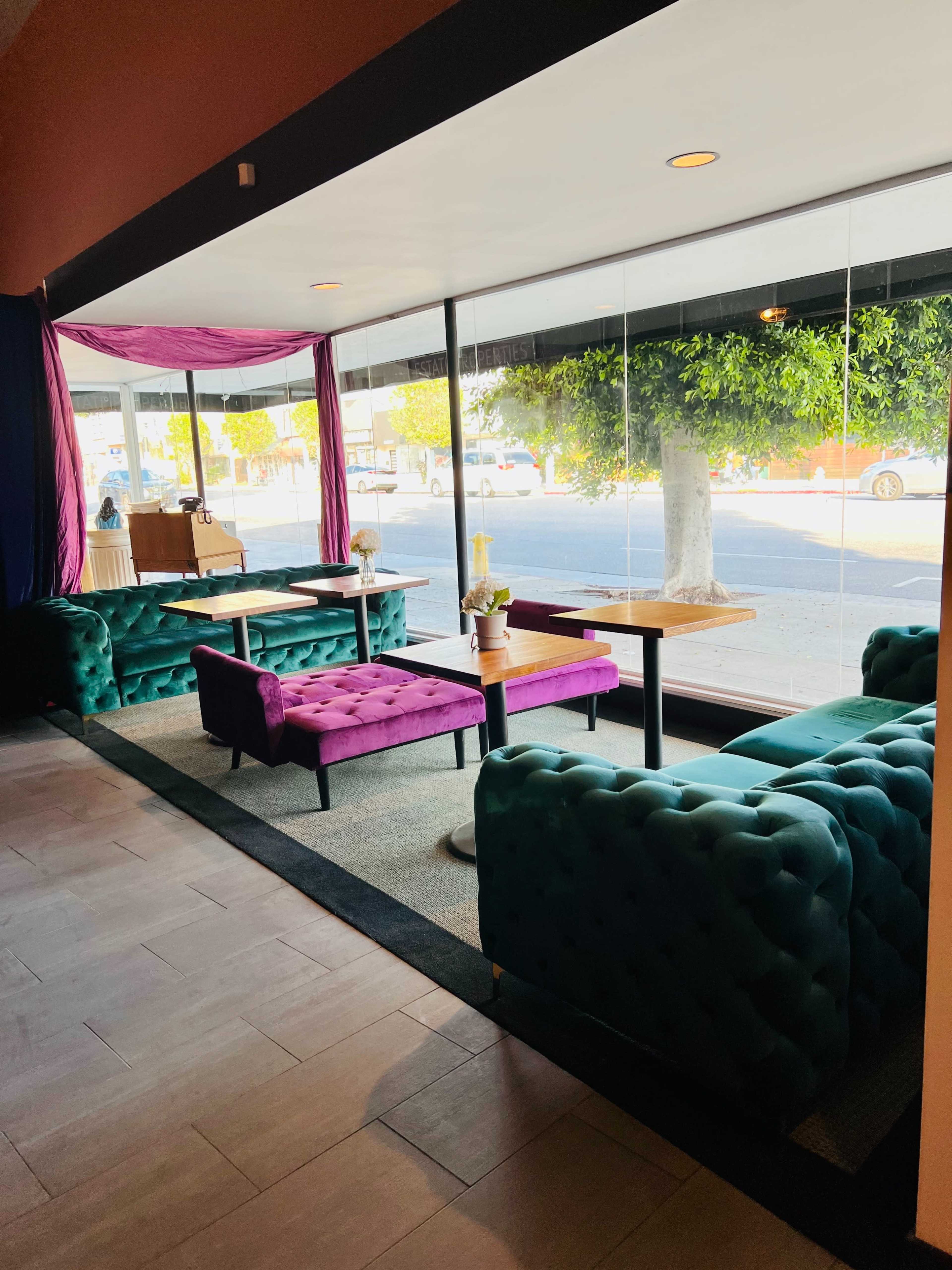 A modern lounge area with tufted teal and purple sofas, contrasting wood tables, and large windows allowing natural light to enter.