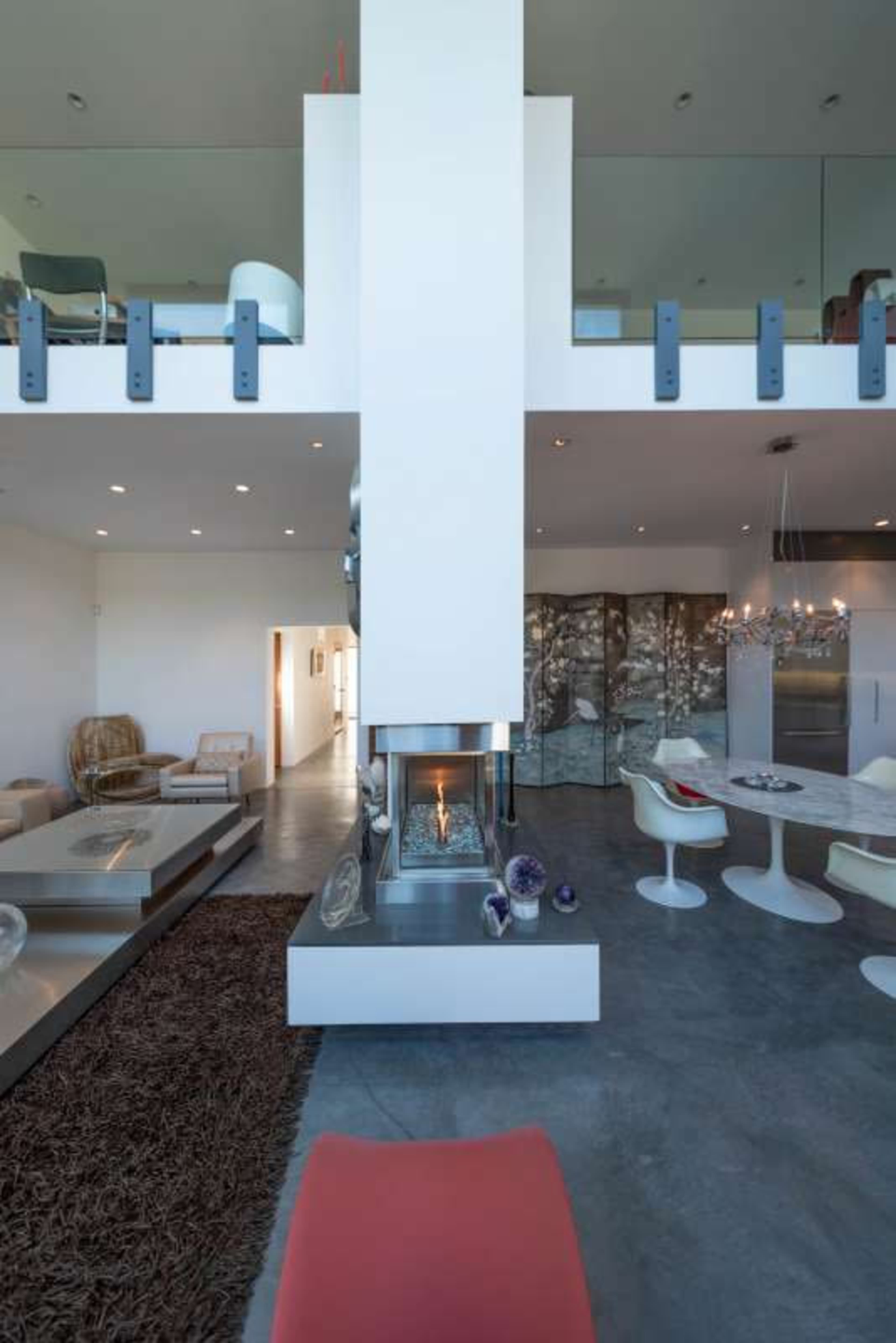 A modern living space featuring a central fireplace, a dining area with sleek chairs, and a second-floor balcony above.