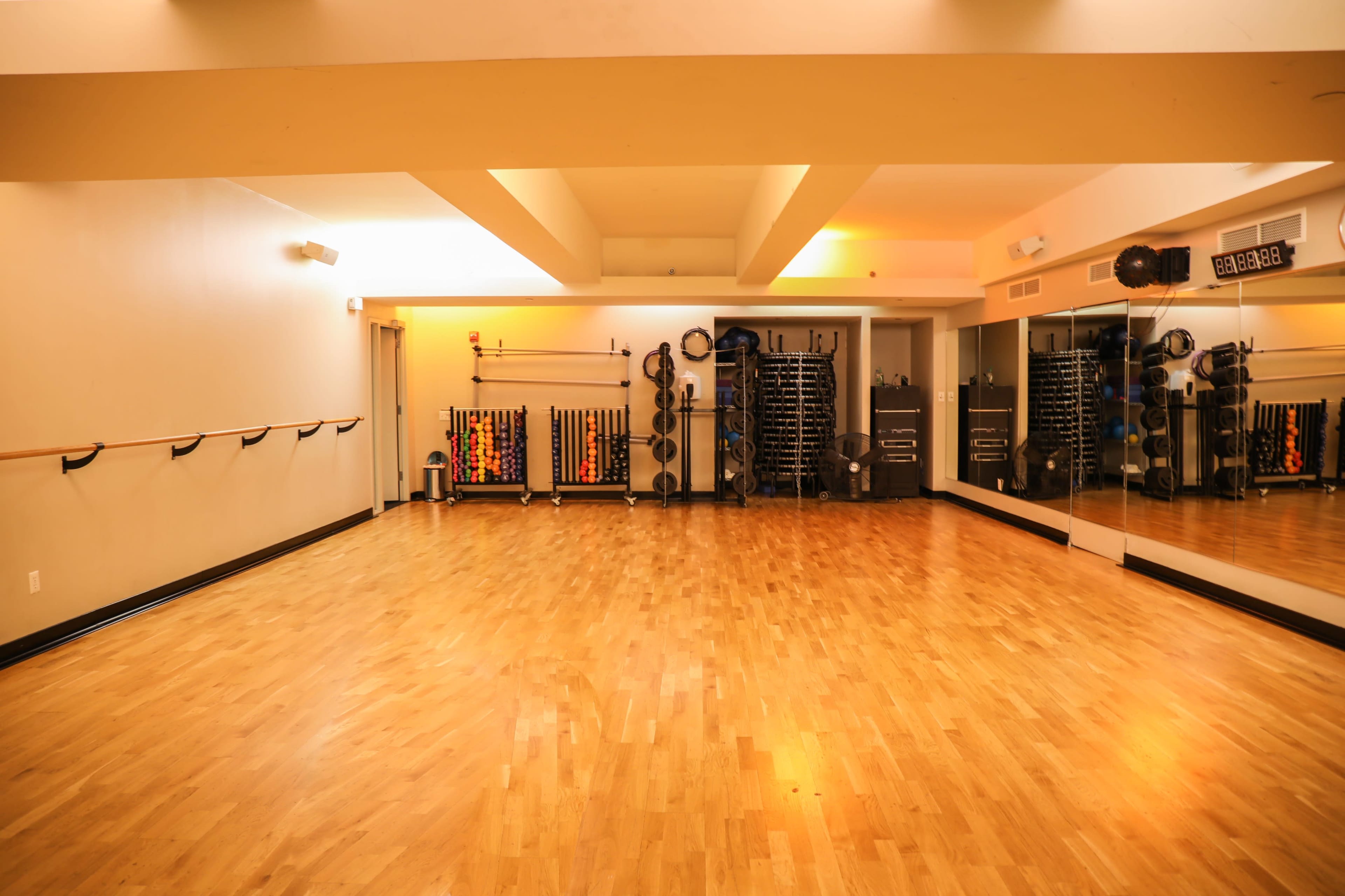 The image shows a spacious fitness studio with wooden flooring, mirrored walls, and various exercise equipment organized on shelves.