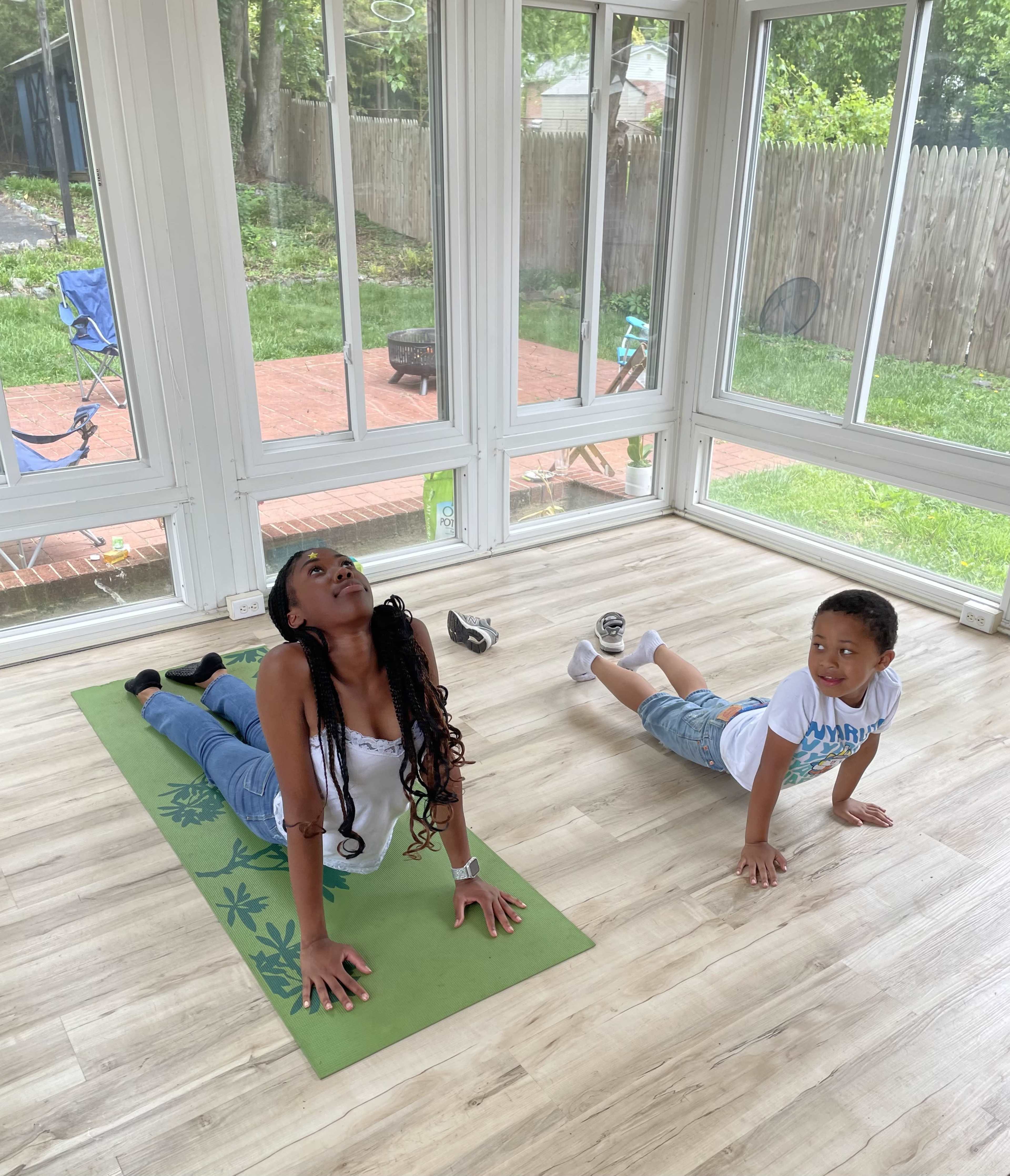 A woman performs a yoga pose on a mat while a child engages in a similar exercise in a sunlit room with large windows.