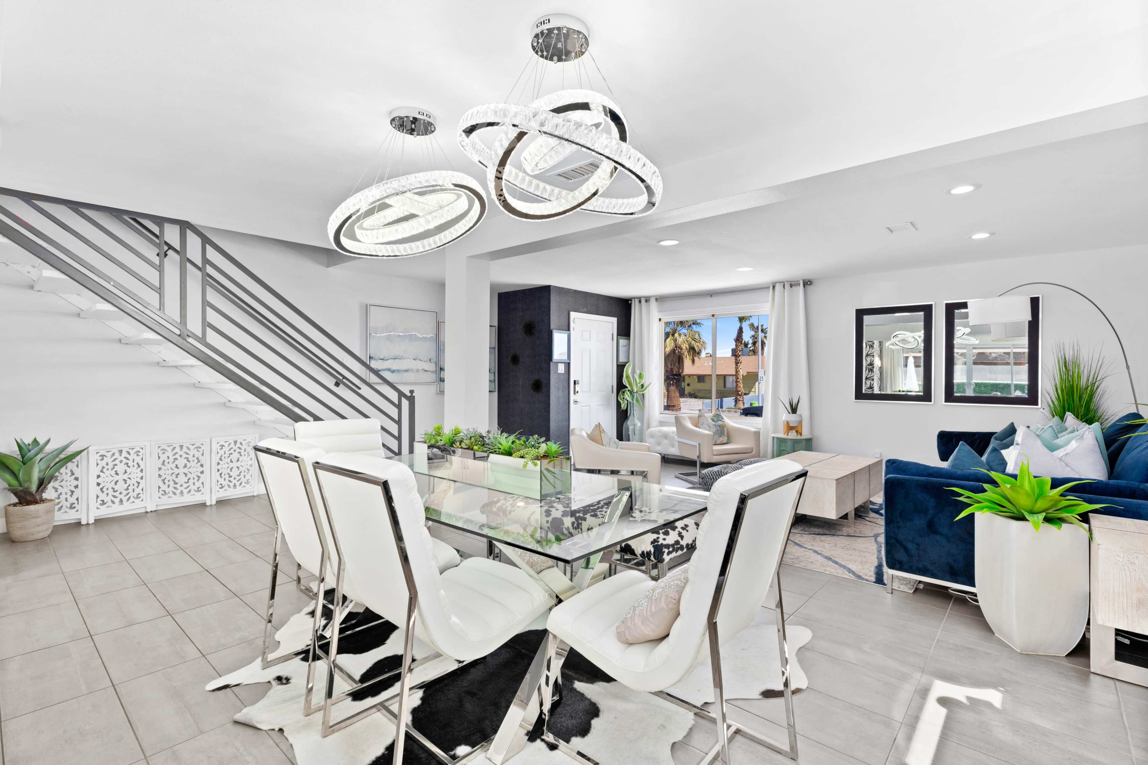 A modern dining area with a glass table and white chairs, adjacent to a living room filled with natural light and decorative plants.