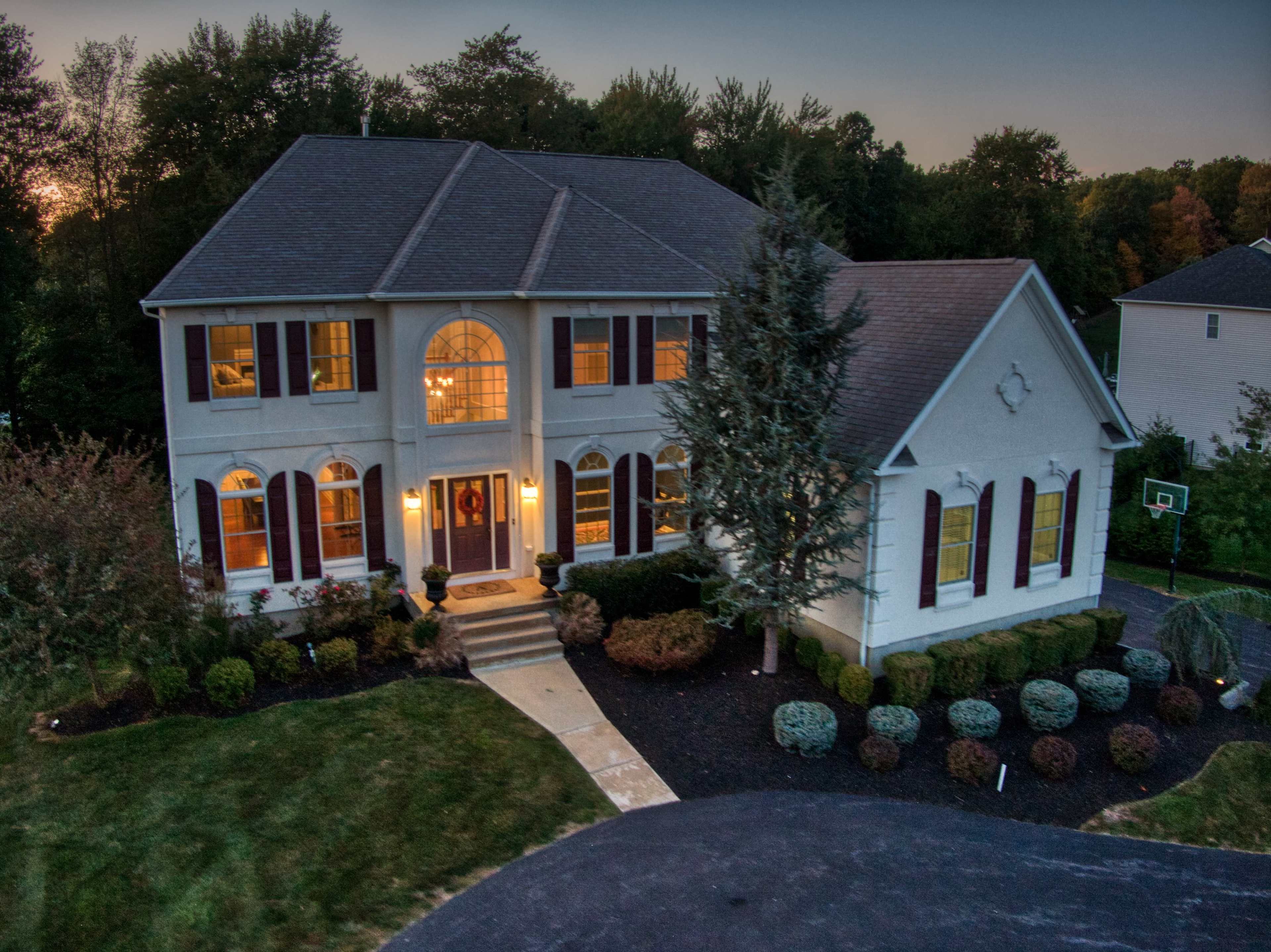 The image shows a large, two-story house with a symmetrical facade, surrounded by landscaped greenery and a stone pathway leading to the front entrance.