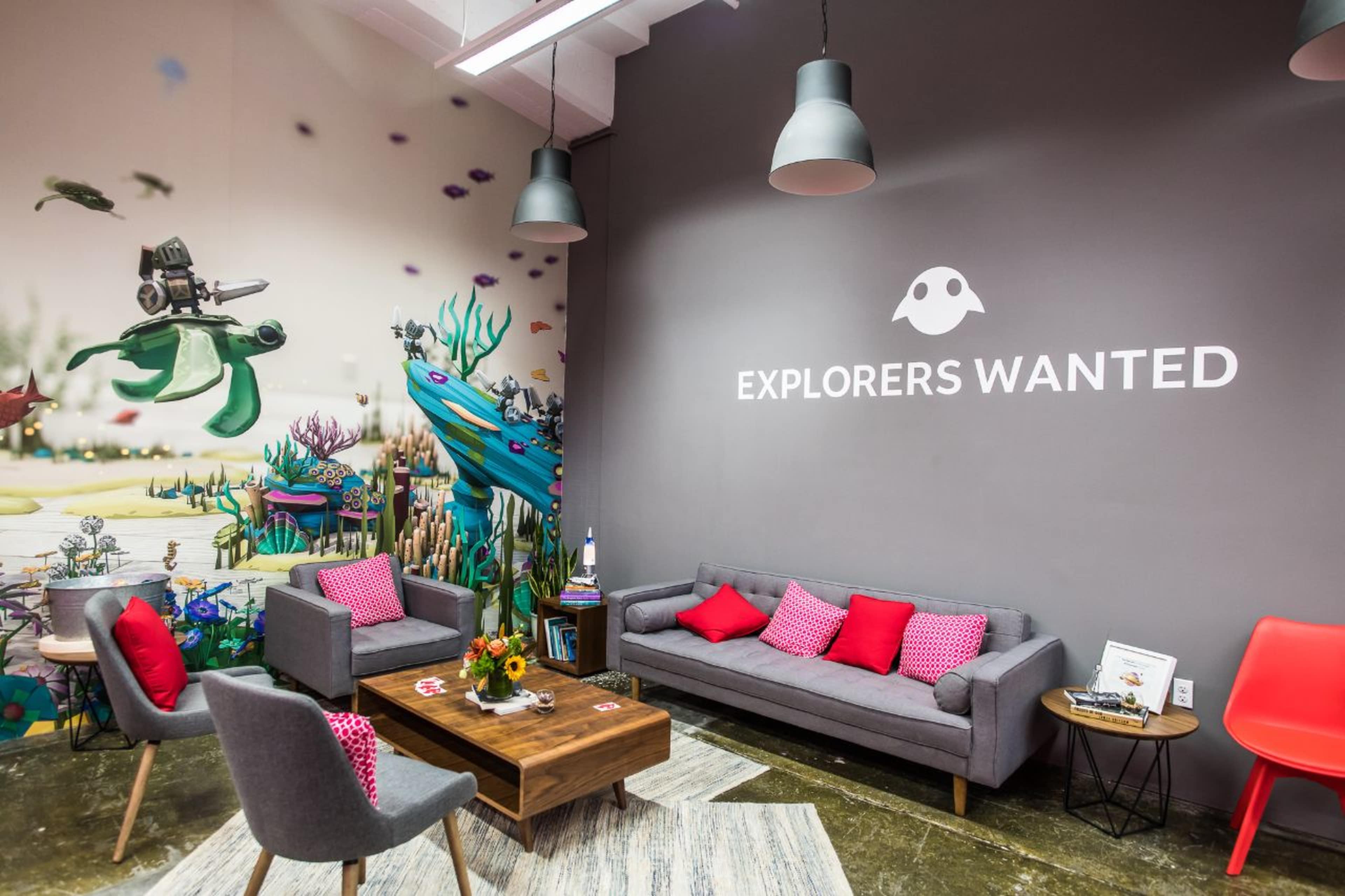 A modern lounge area with gray sofas, a wooden coffee table, and a colorful mural featuring aquatic themes on one wall, along with the phrase "EXPLORERS WANTED" displayed prominently.