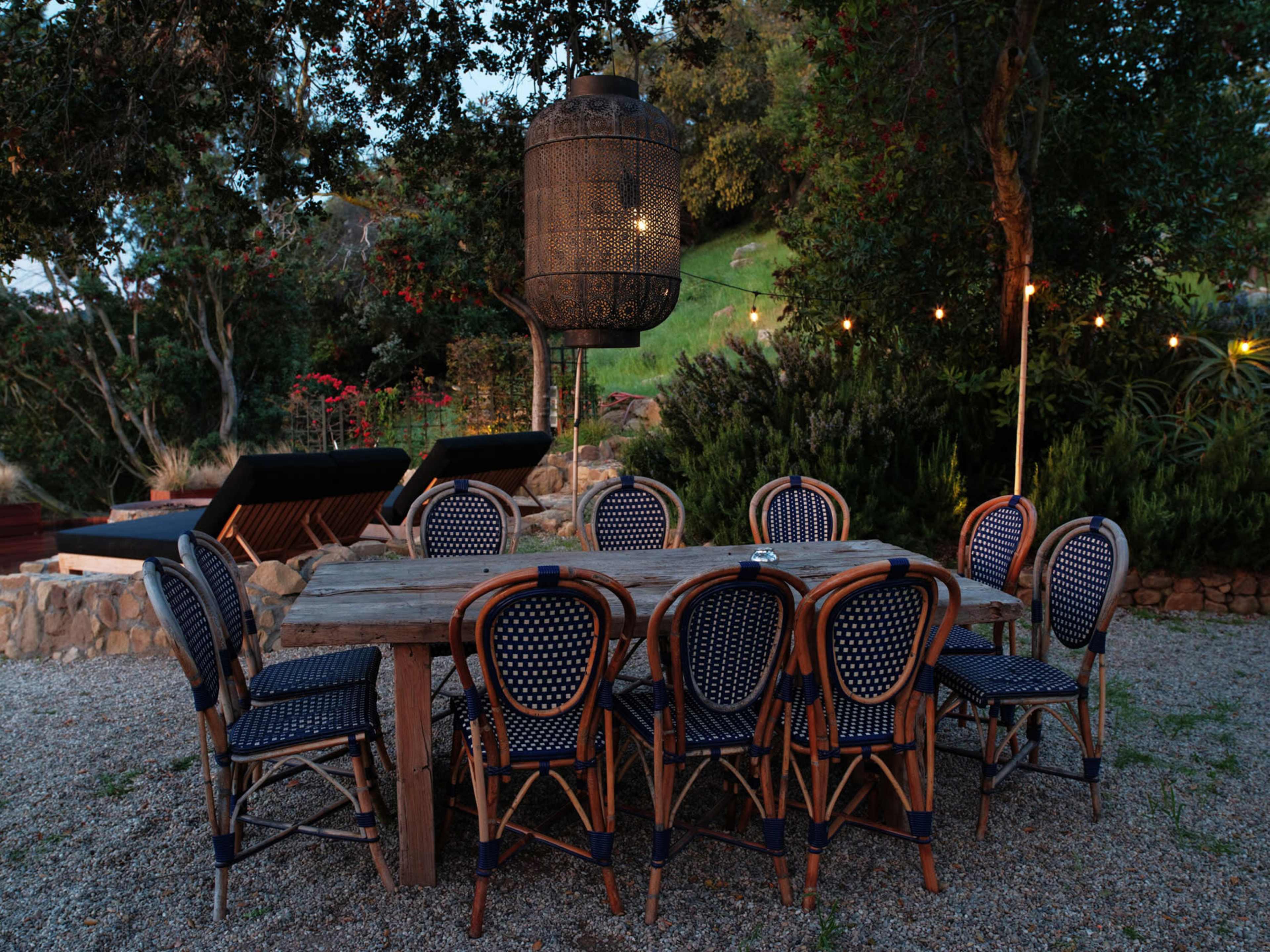 A large wooden dining table surrounded by ten wicker chairs is set in a gravel area, with a decorative lantern hanging above and string lights illuminating the surrounding garden.