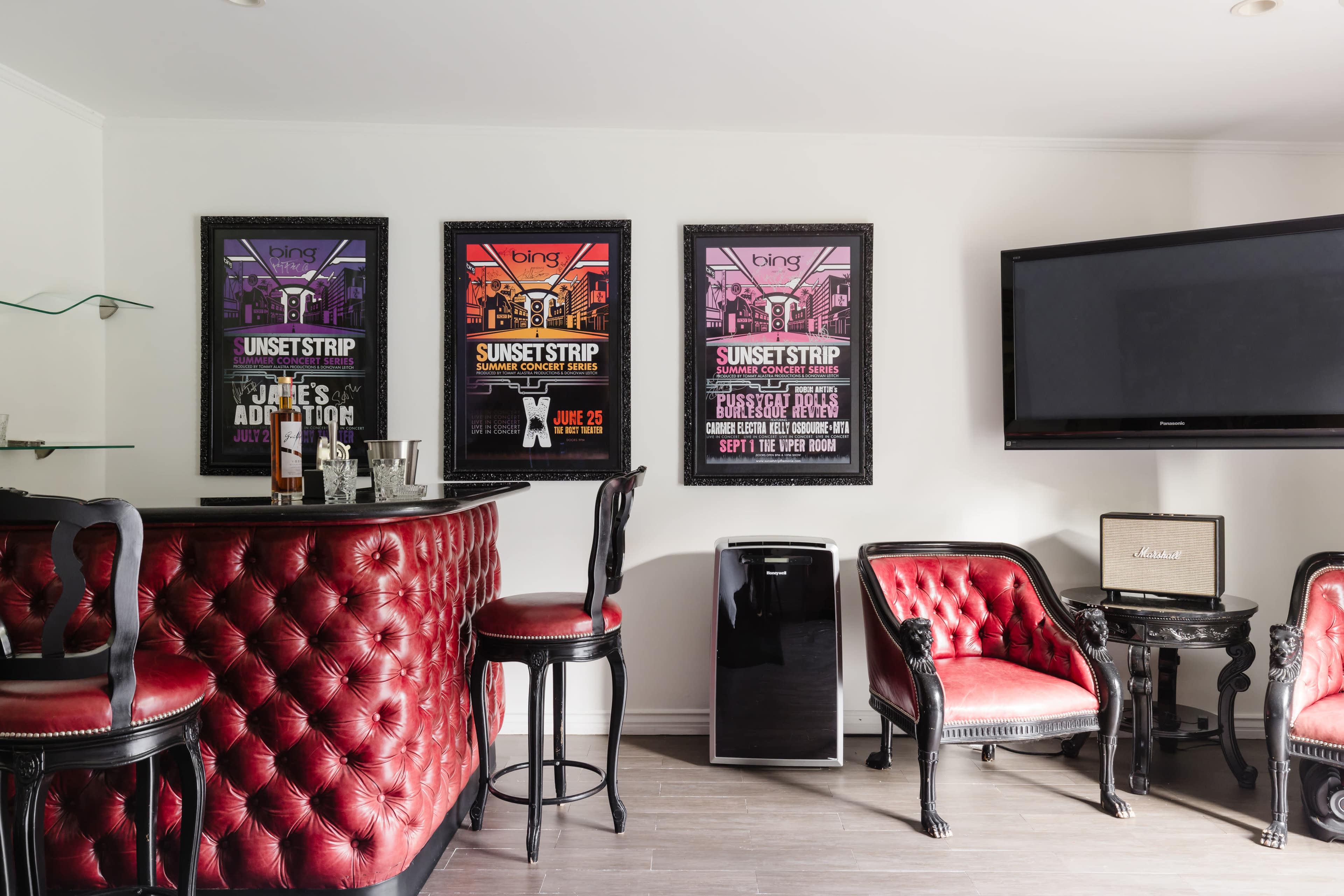 A stylish lounge area featuring a bar with red tufted upholstery, framed colorful event posters on the wall, and elegant black furniture.
