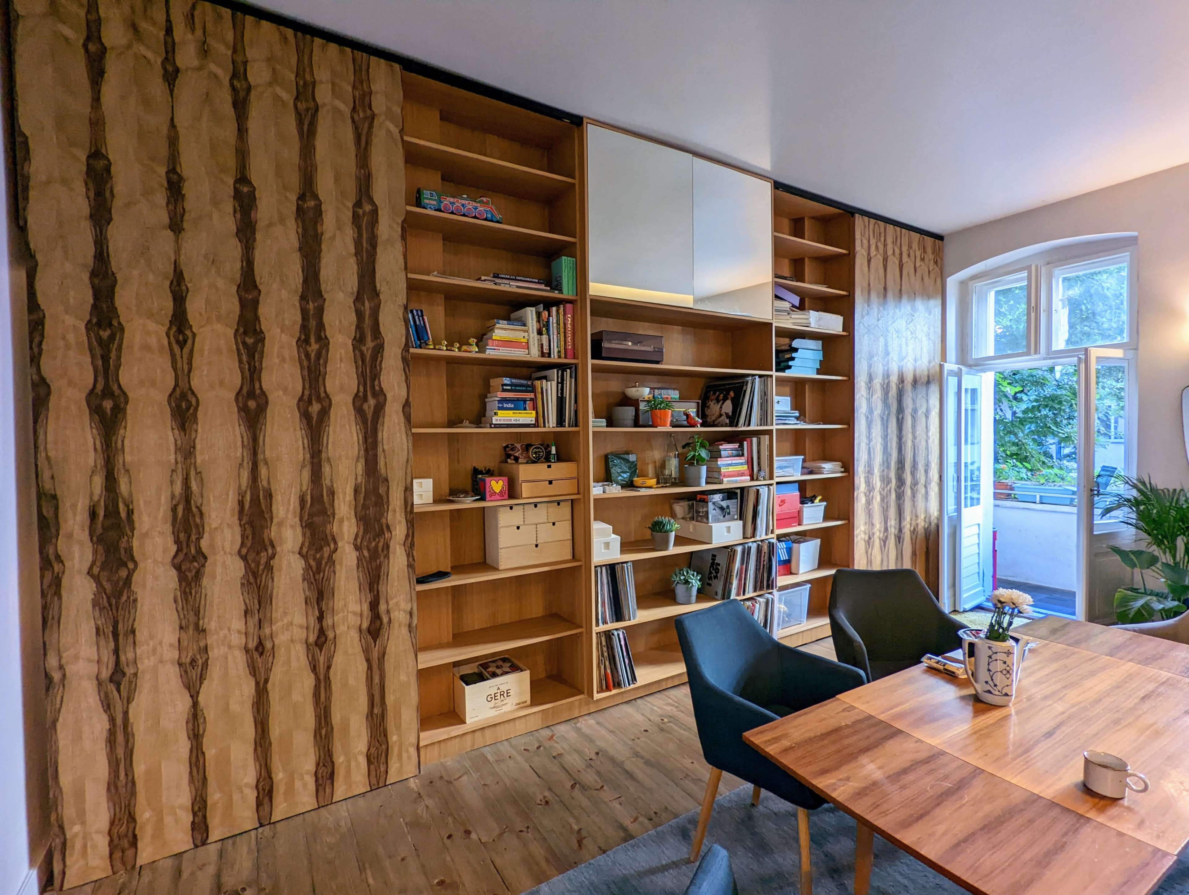 A modern interior with a wooden bookshelf against one wall, a dining table set with a vase, and large windows letting in natural light.