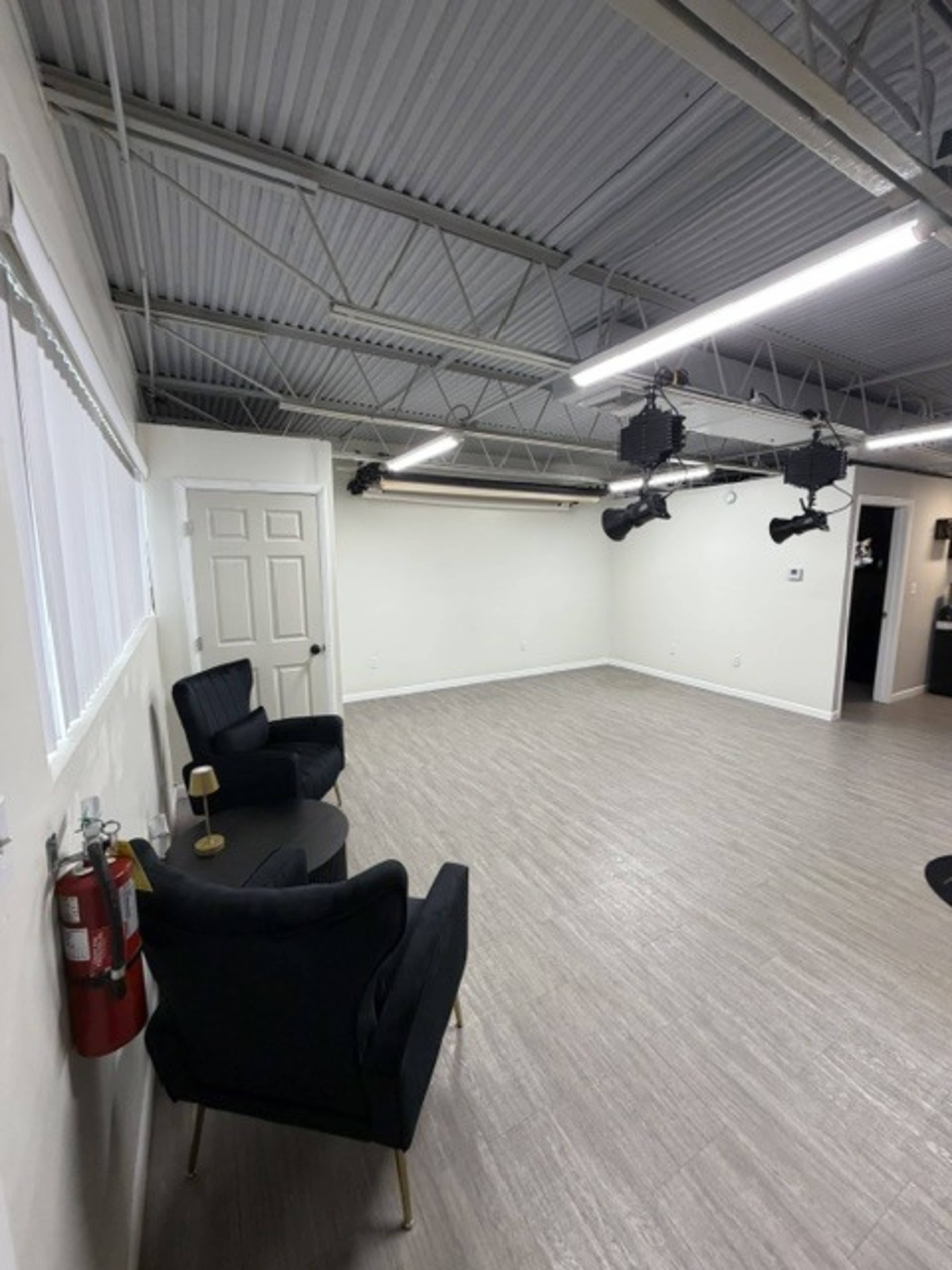 The image shows a spacious, empty room with hardwood flooring, two black armchairs, a small table, and overhead lighting fixtures.