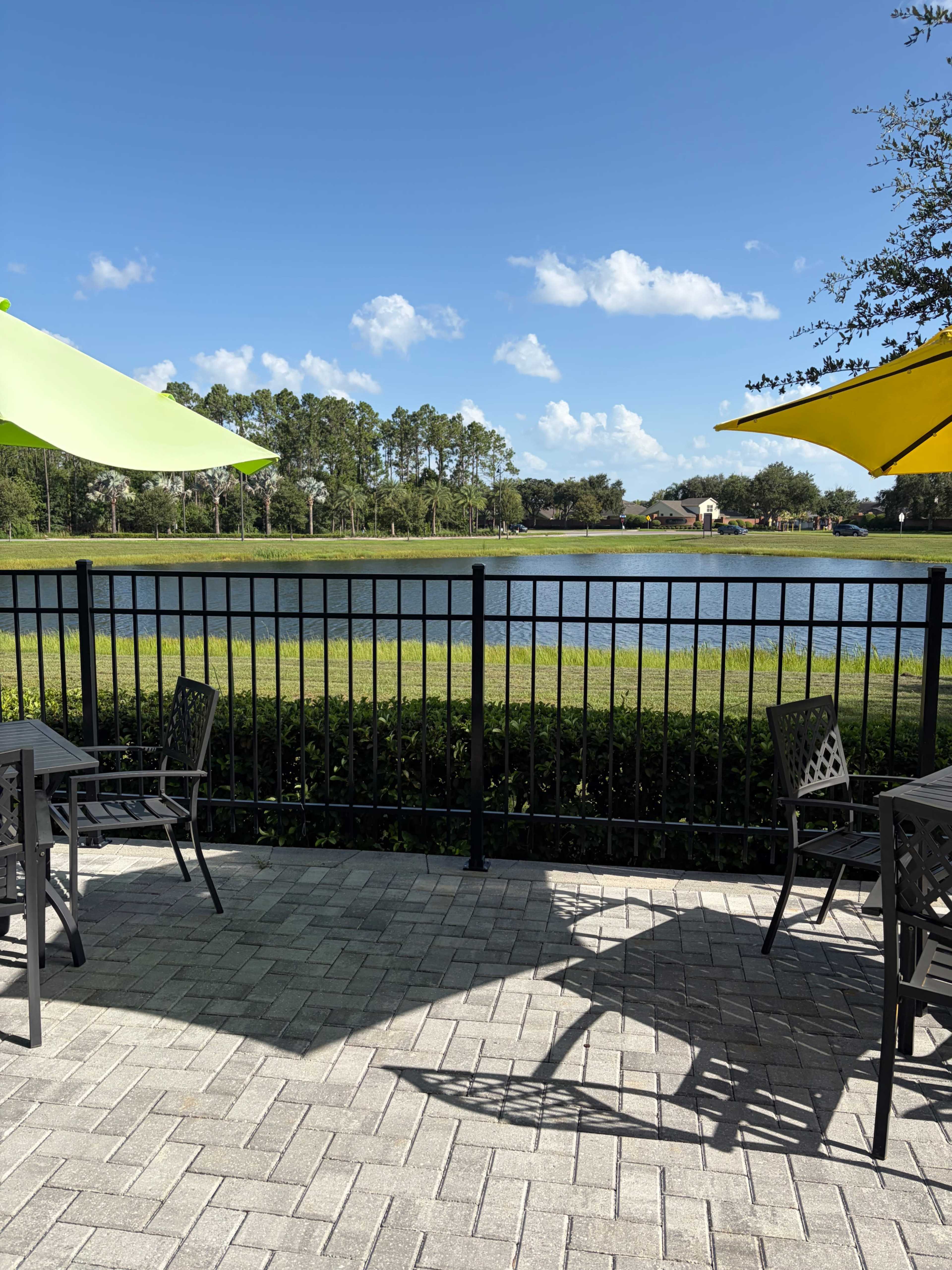 A patio area with metal tables and chairs overlooks a serene lake surrounded by trees under a bright blue sky with a few clouds.