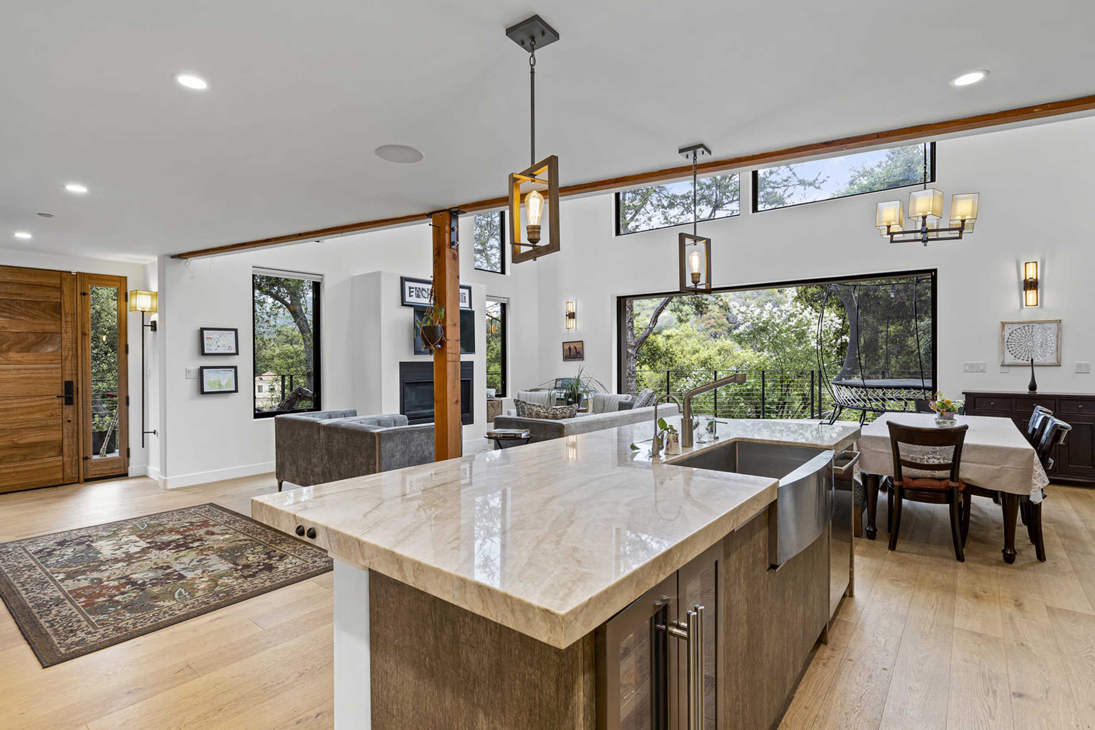 The image shows a modern open-concept living space featuring a kitchen with a large island, a dining area with a table, and a comfortable seating area with large windows overlooking greenery.