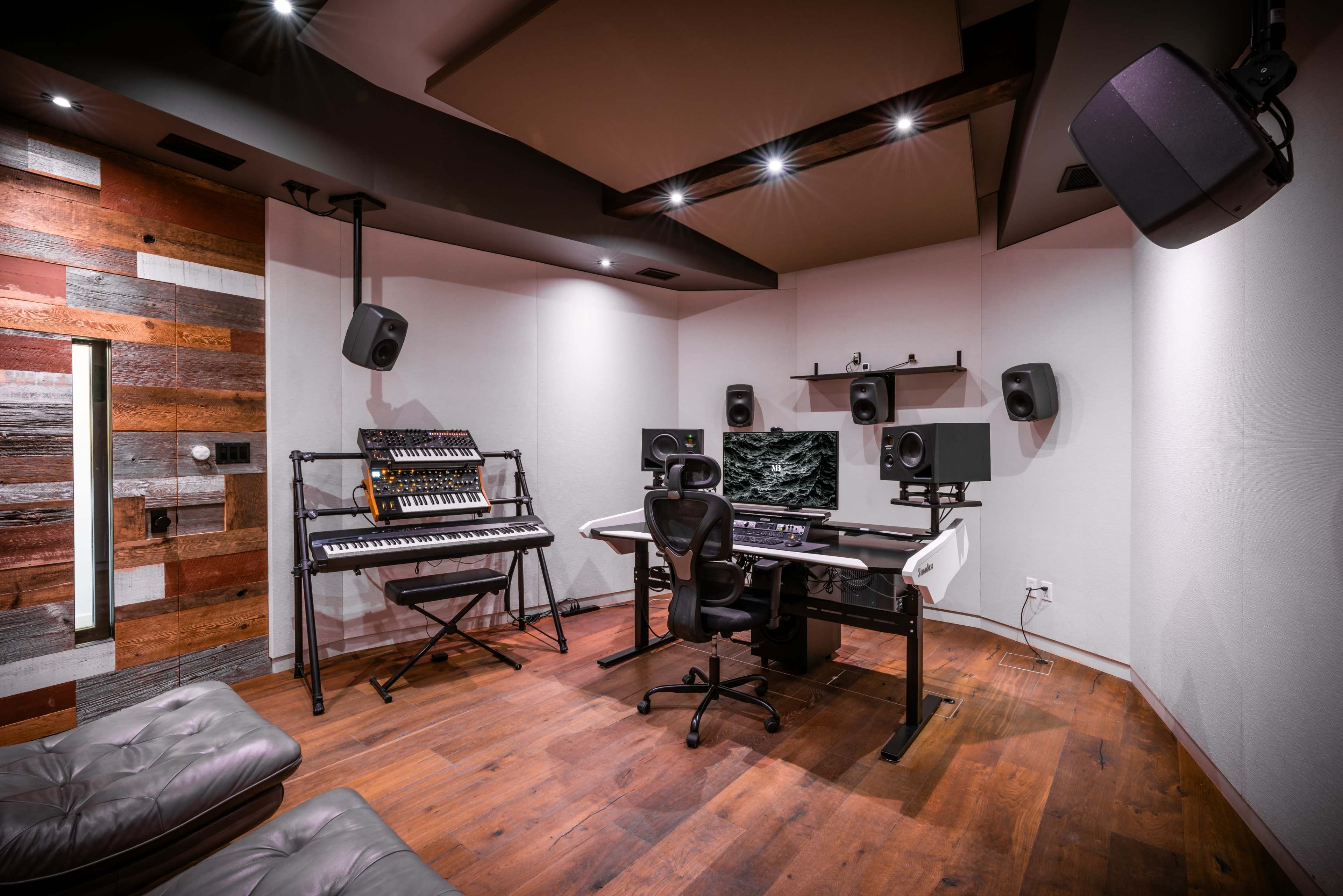 The image shows a modern music studio featuring a keyboard station, a large desk with a computer and audio equipment, and sound monitors mounted on the walls.