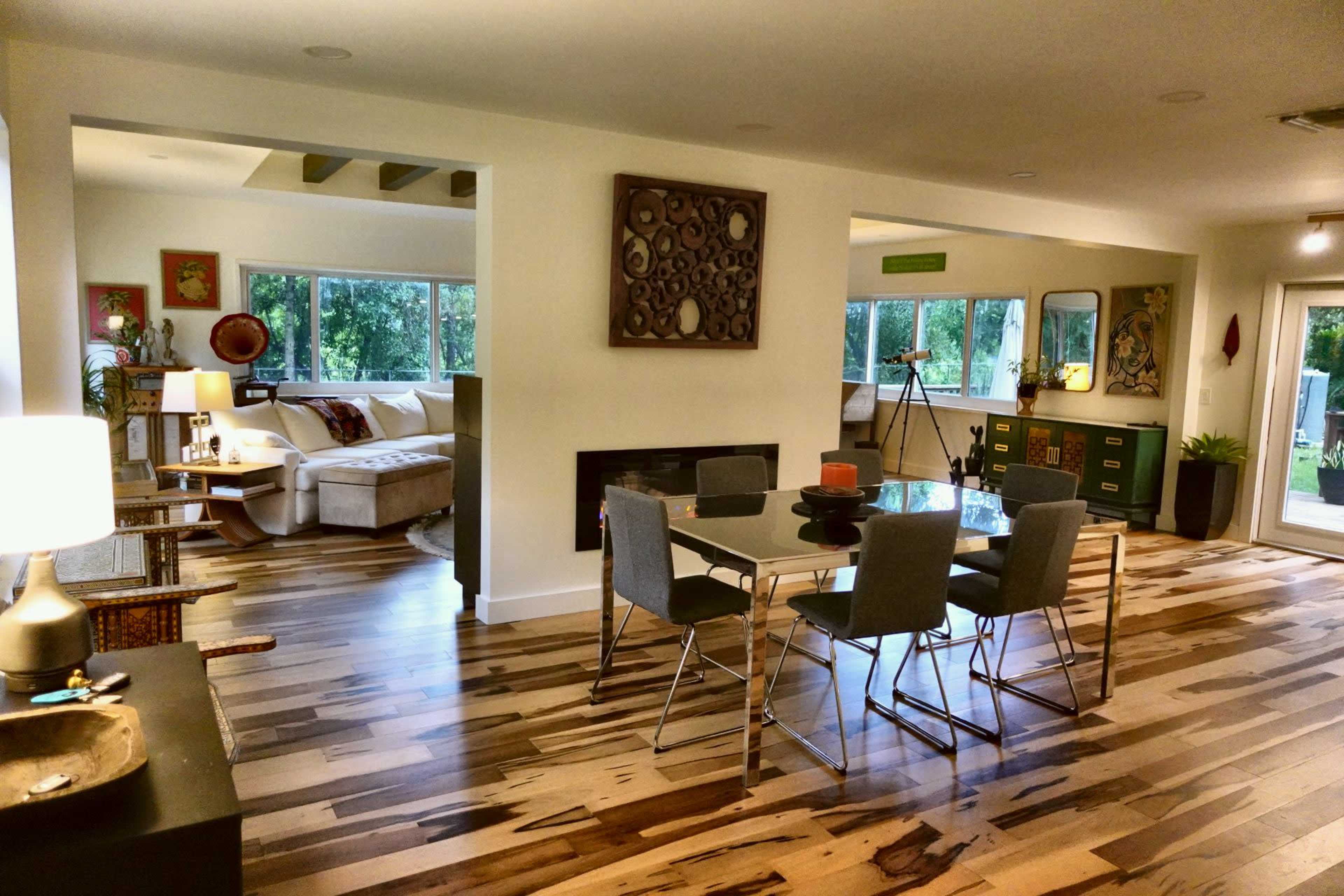 A modern dining area features a glass table surrounded by gray chairs, adjacent to a living space with a sectional sofa and large windows.