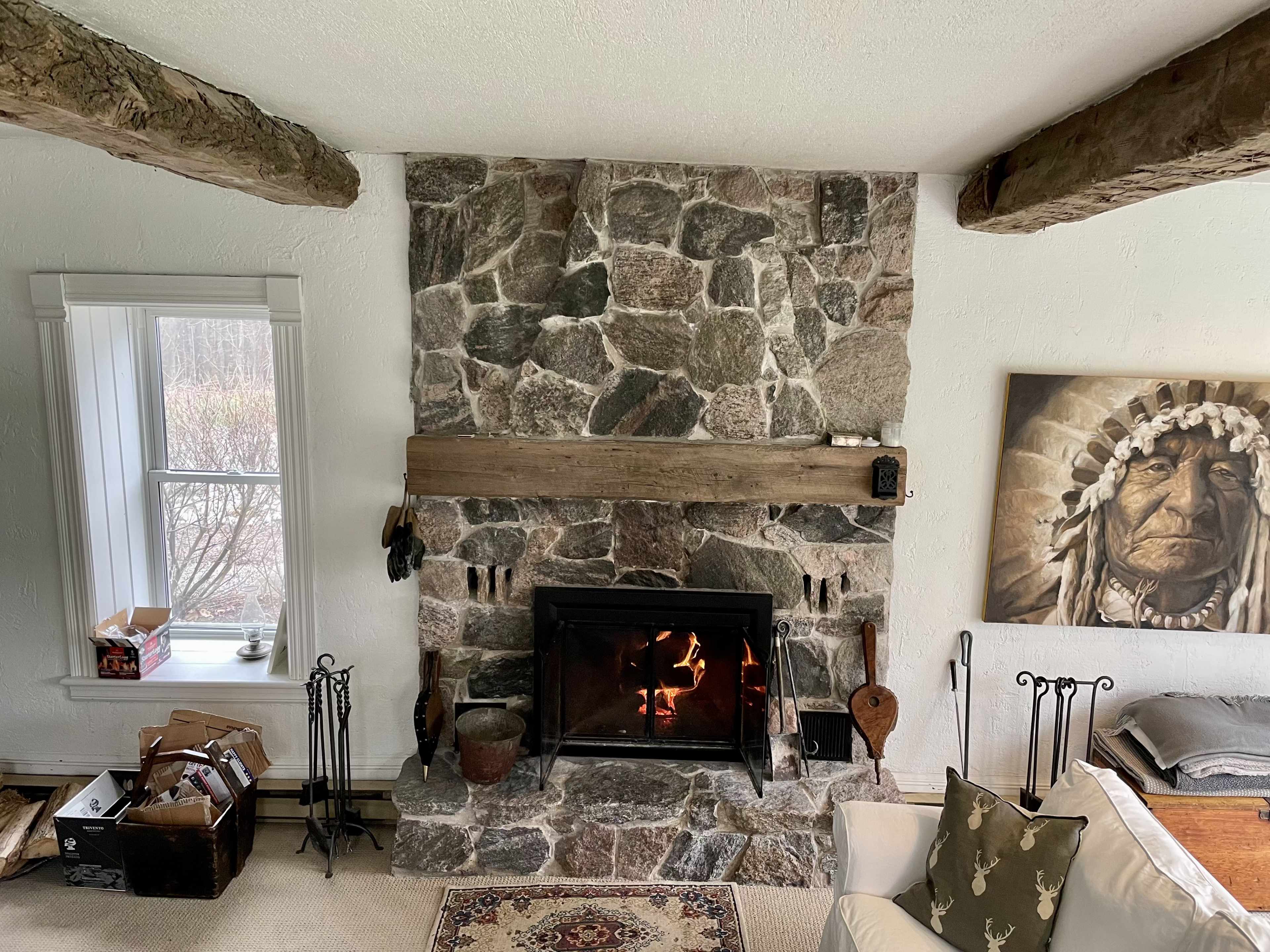 A living room with a stone fireplace, wooden beams overhead, and a window beside a box on the floor.