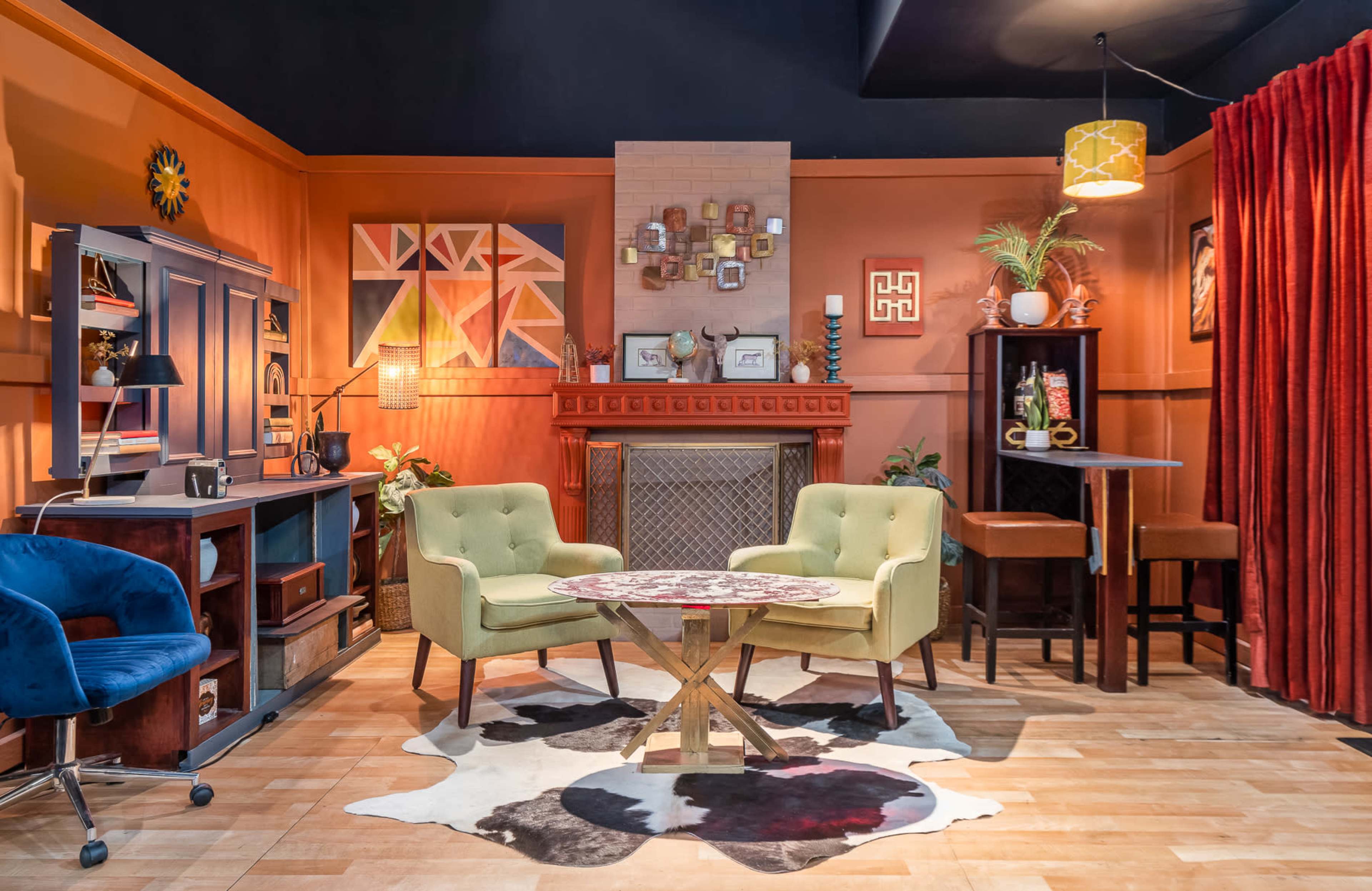A cozy room featuring a fireplace with decorative elements, two green chairs, a wooden coffee table, and various artworks on the walls.