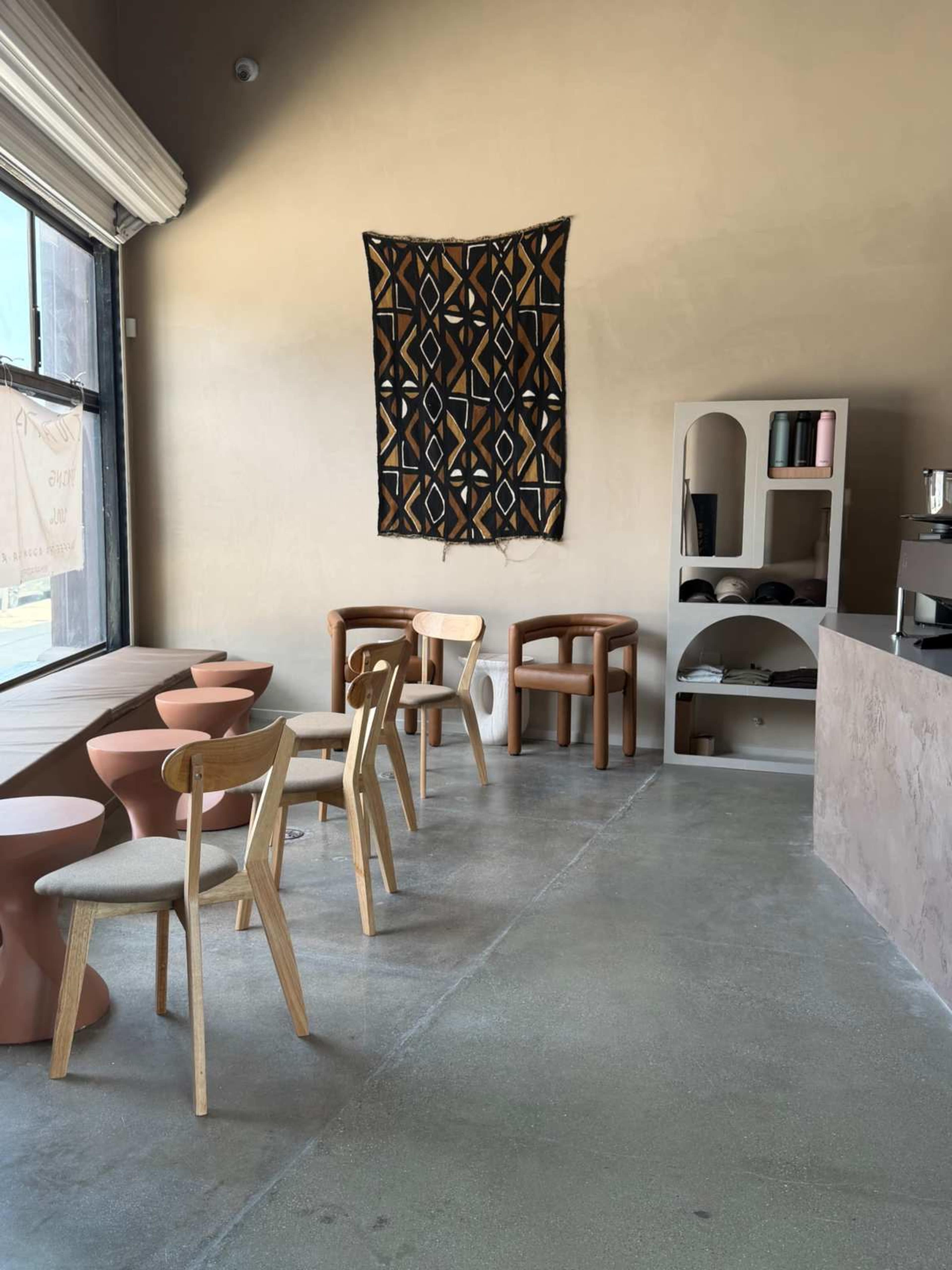 The image shows a modern café interior featuring a row of wooden chairs, small circular tables, a patterned wall hanging, and a minimalist shelving unit.