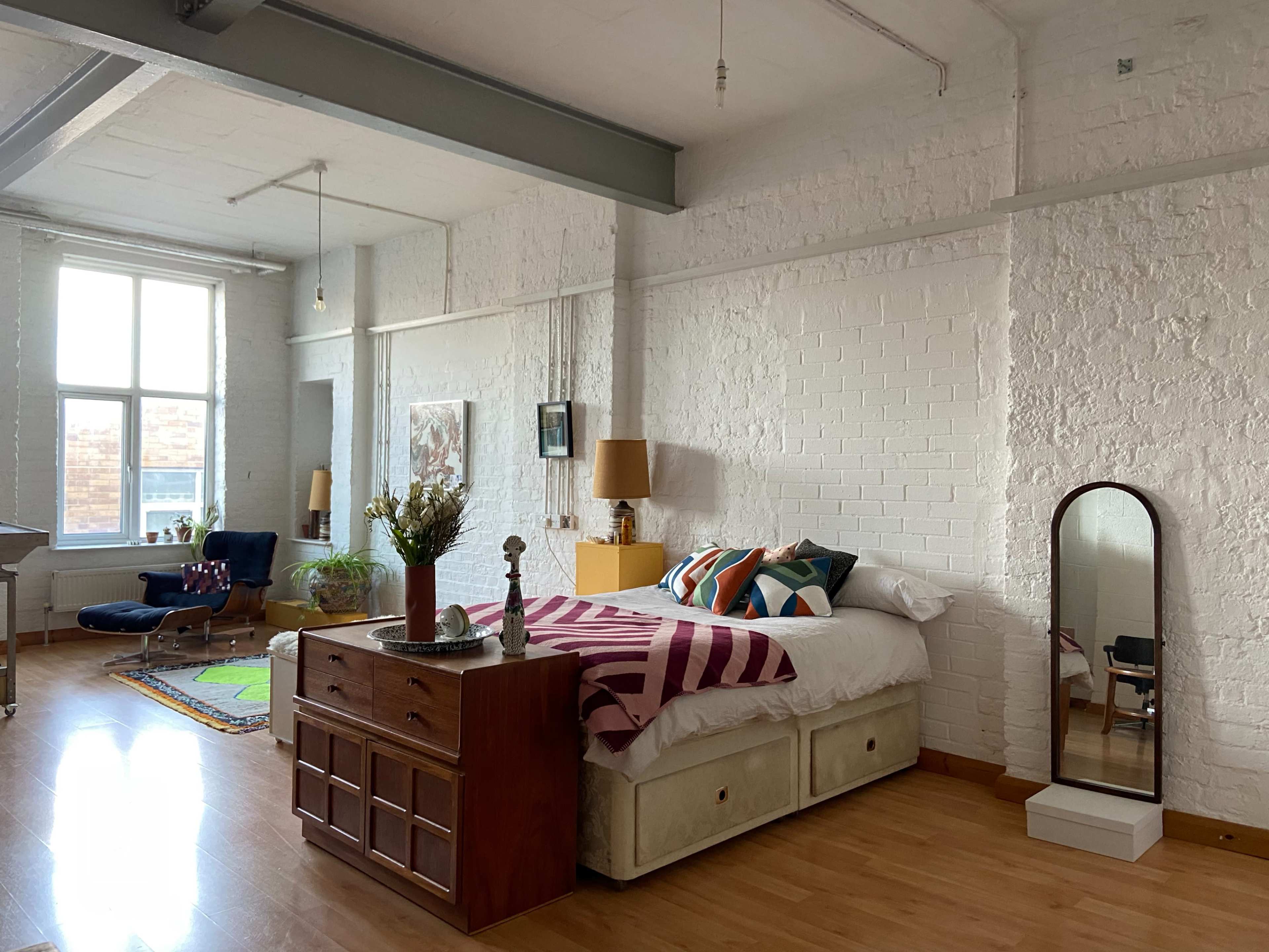 A bright, spacious room with a bed, a wooden dresser, a large mirror, and a colorful area rug.