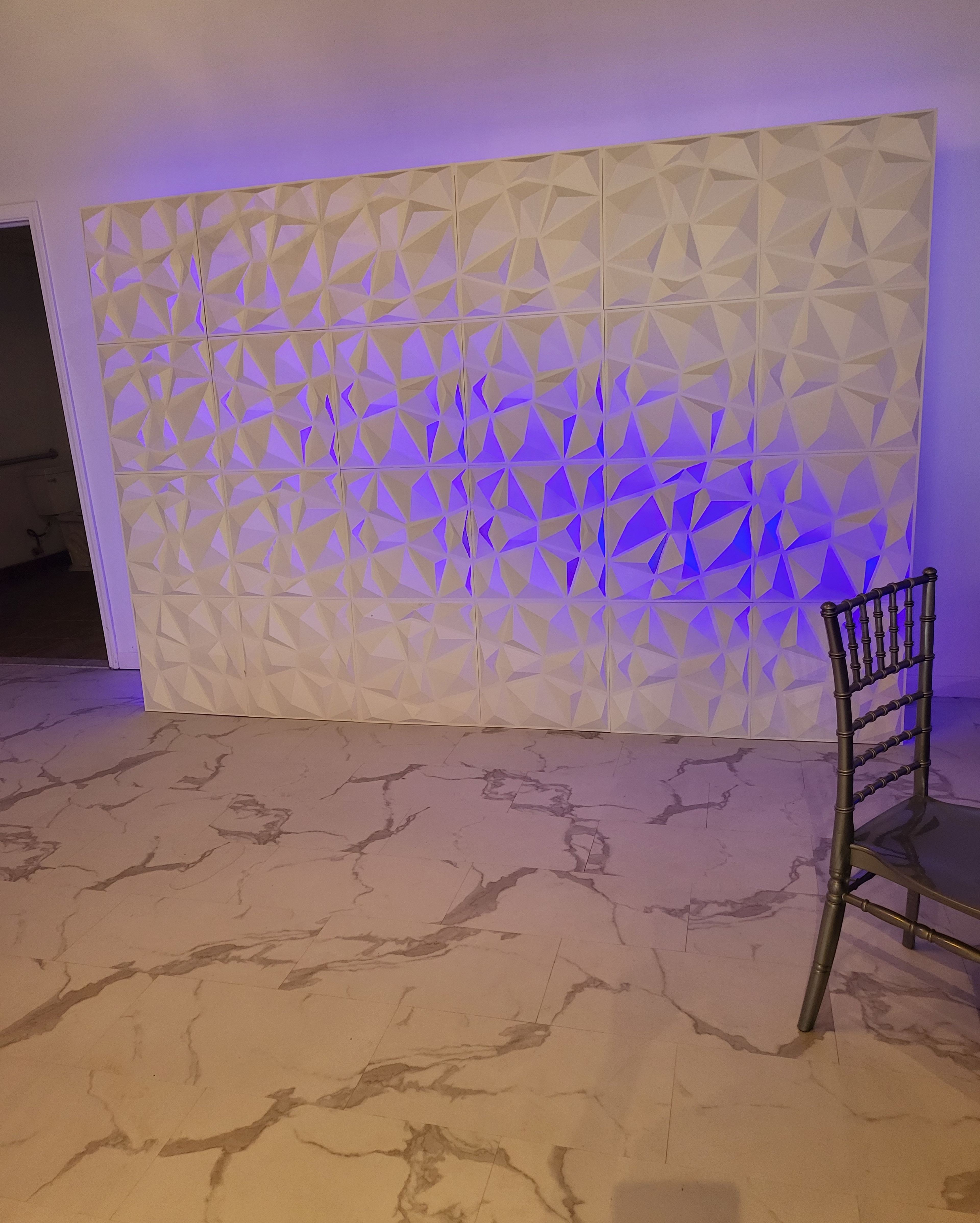 A patterned wall with triangular designs illuminated by blue light, alongside a single gray chair on a marble floor.