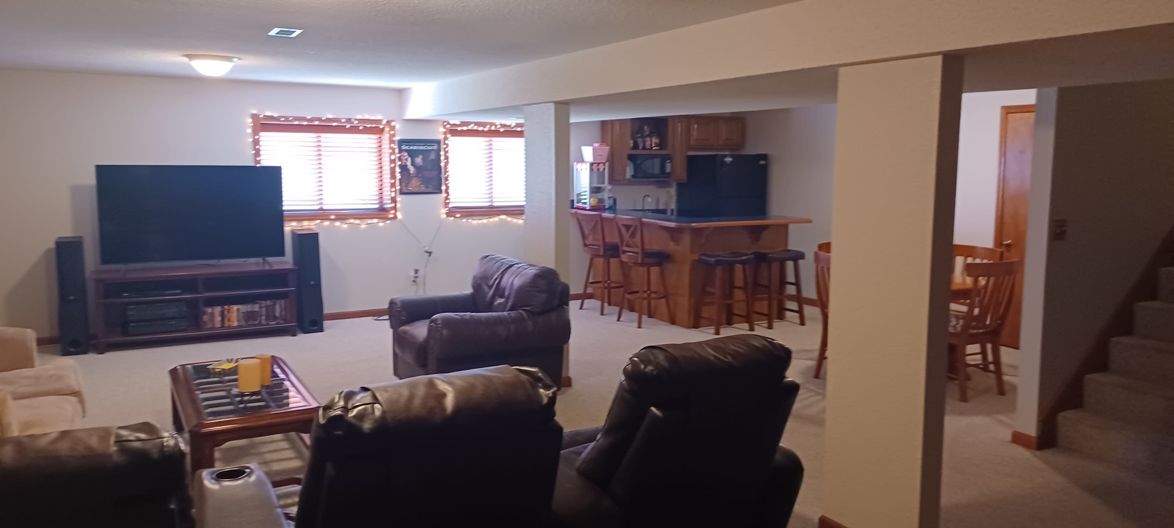 The image shows a finished basement living area with a television, seating, a kitchenette bar, and a staircase leading to another level.
