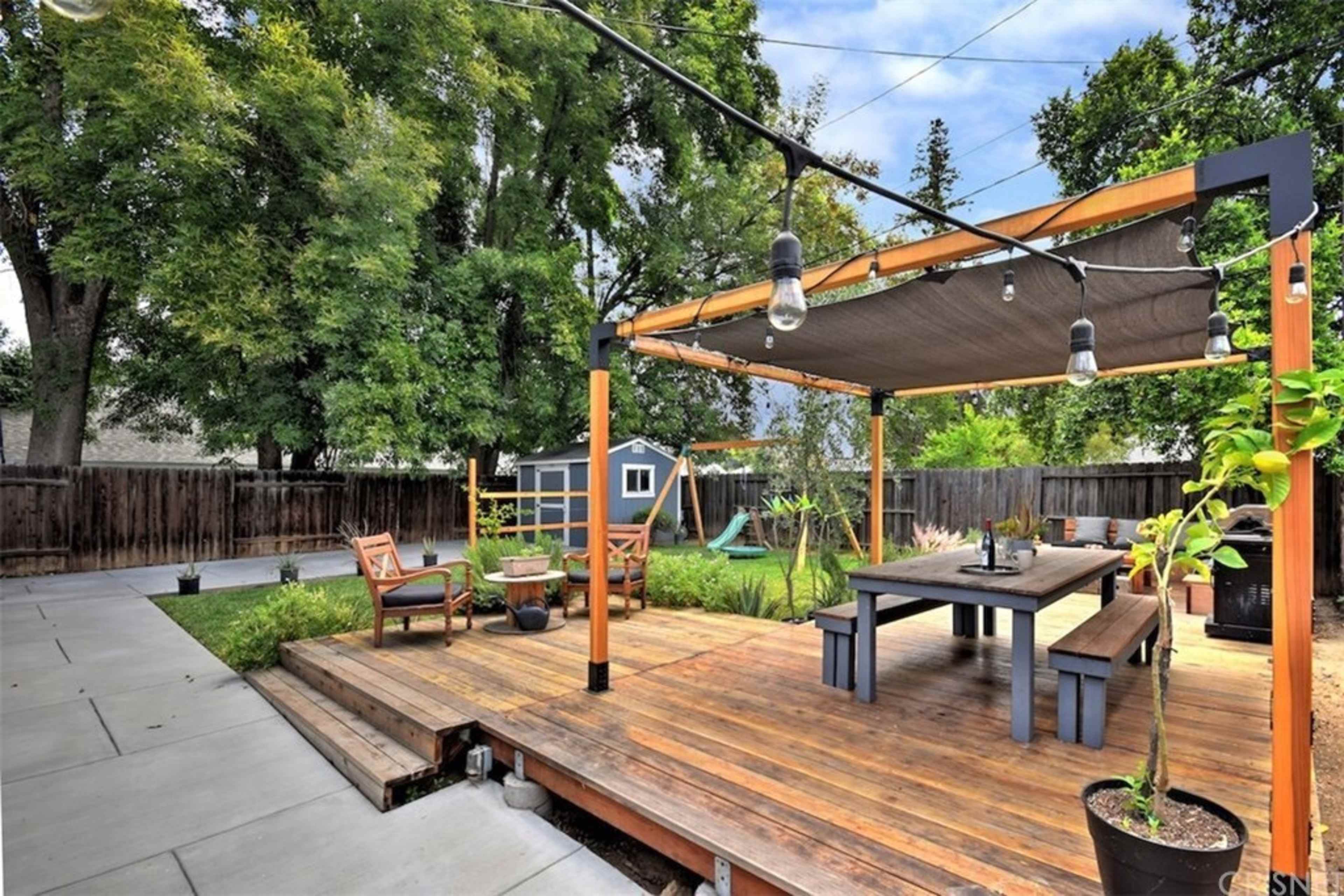 The image shows a wooden patio with a dining table and lounge chairs, surrounded by greenery and a shed in the background.