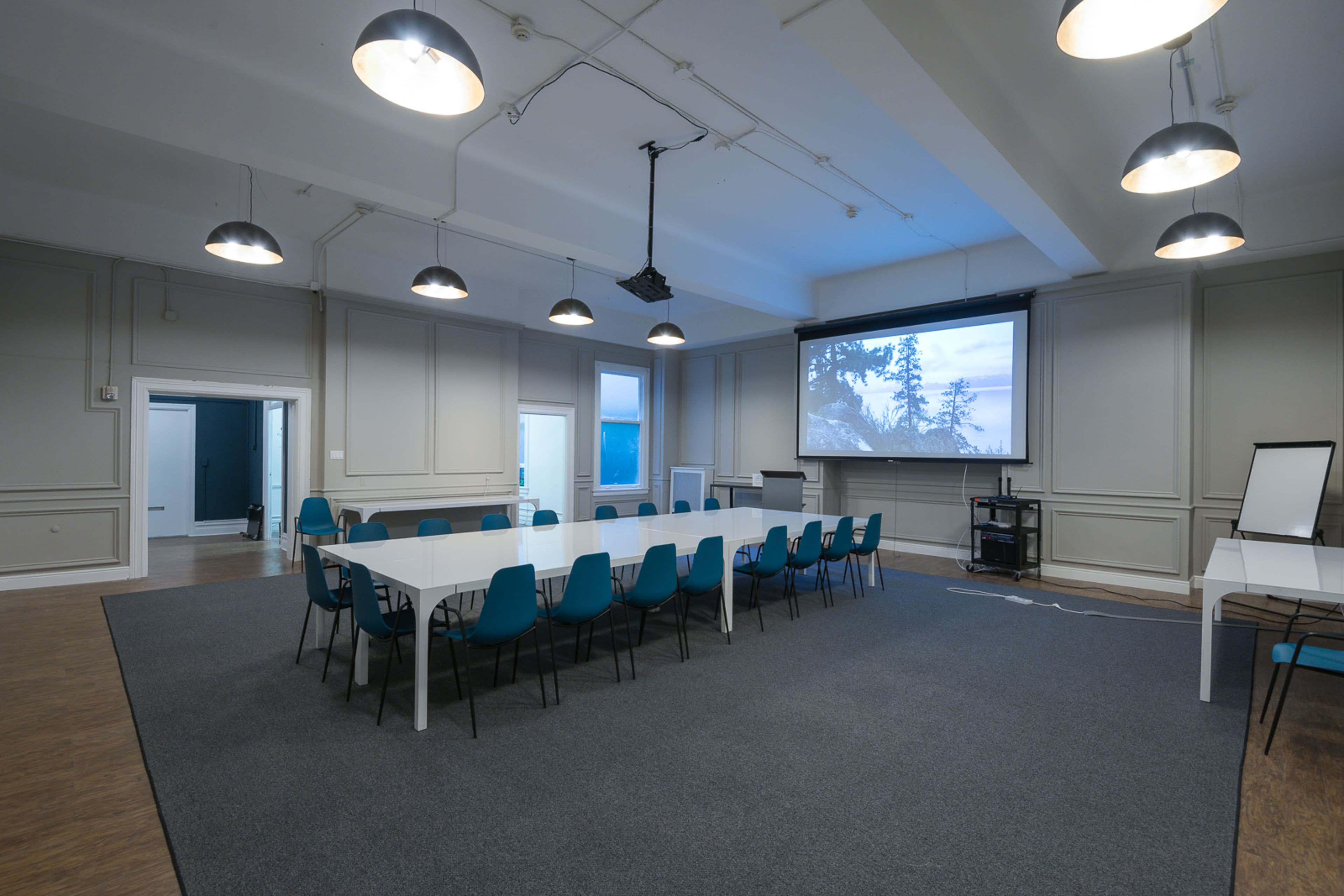 A spacious conference room features a long white table surrounded by teal chairs, with a projector screen mounted on the wall and track lighting overhead.