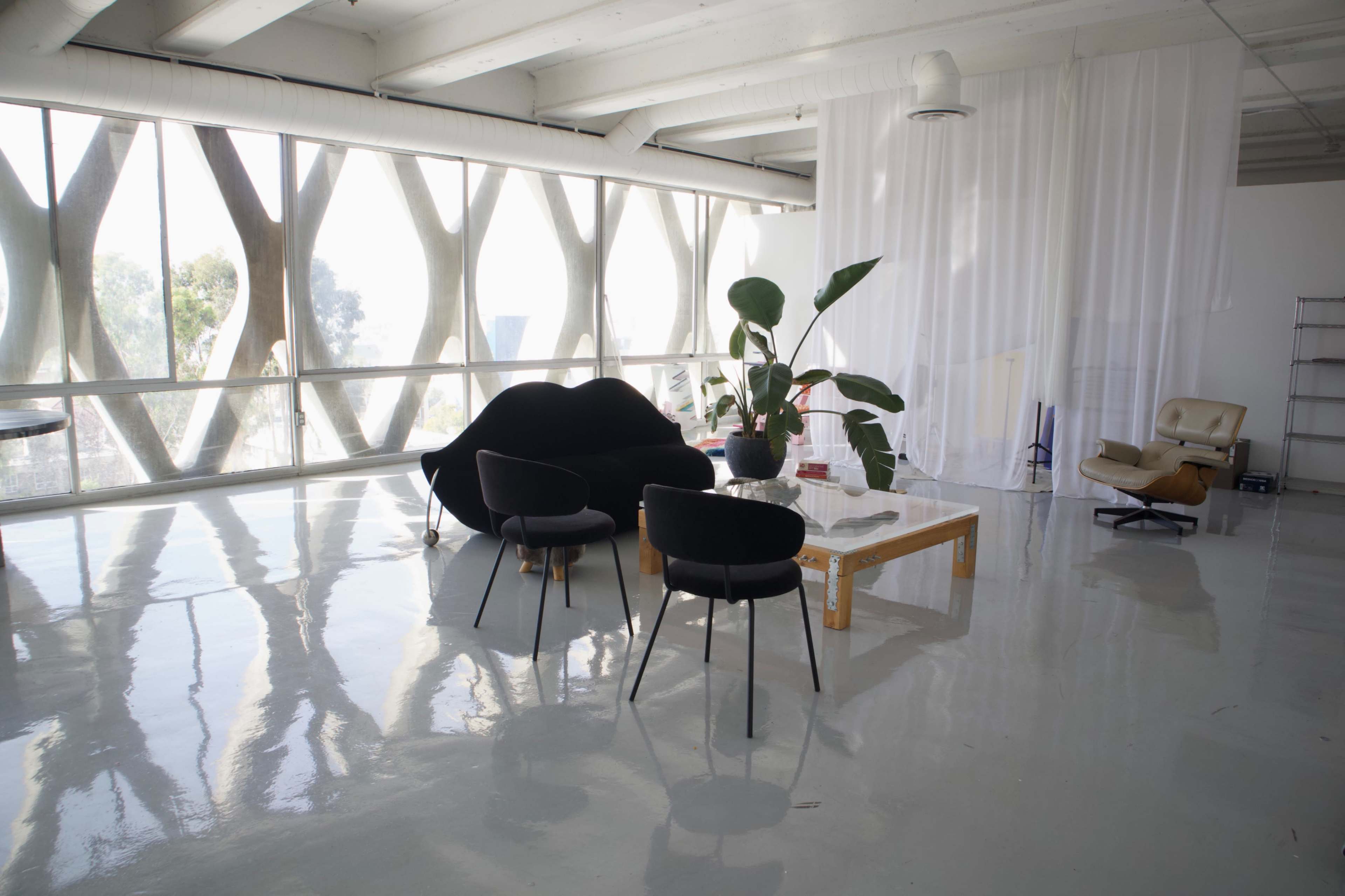 A modern interior space features large windows, a black sofa, two black chairs, a wooden coffee table, and a potted plant.