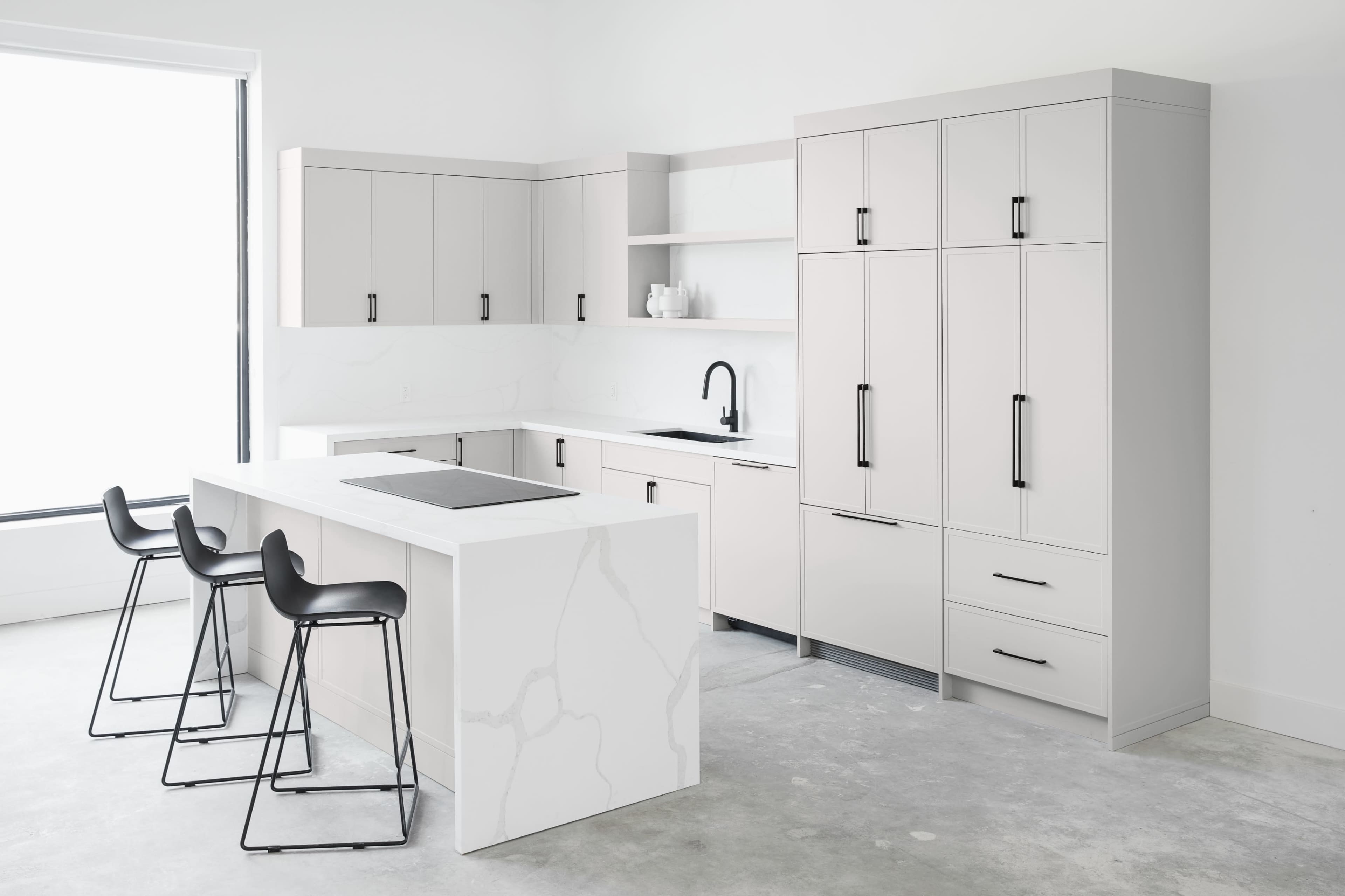 A modern kitchen features minimalist cabinetry, a large island with seating, and sleek appliances against a light-colored background.