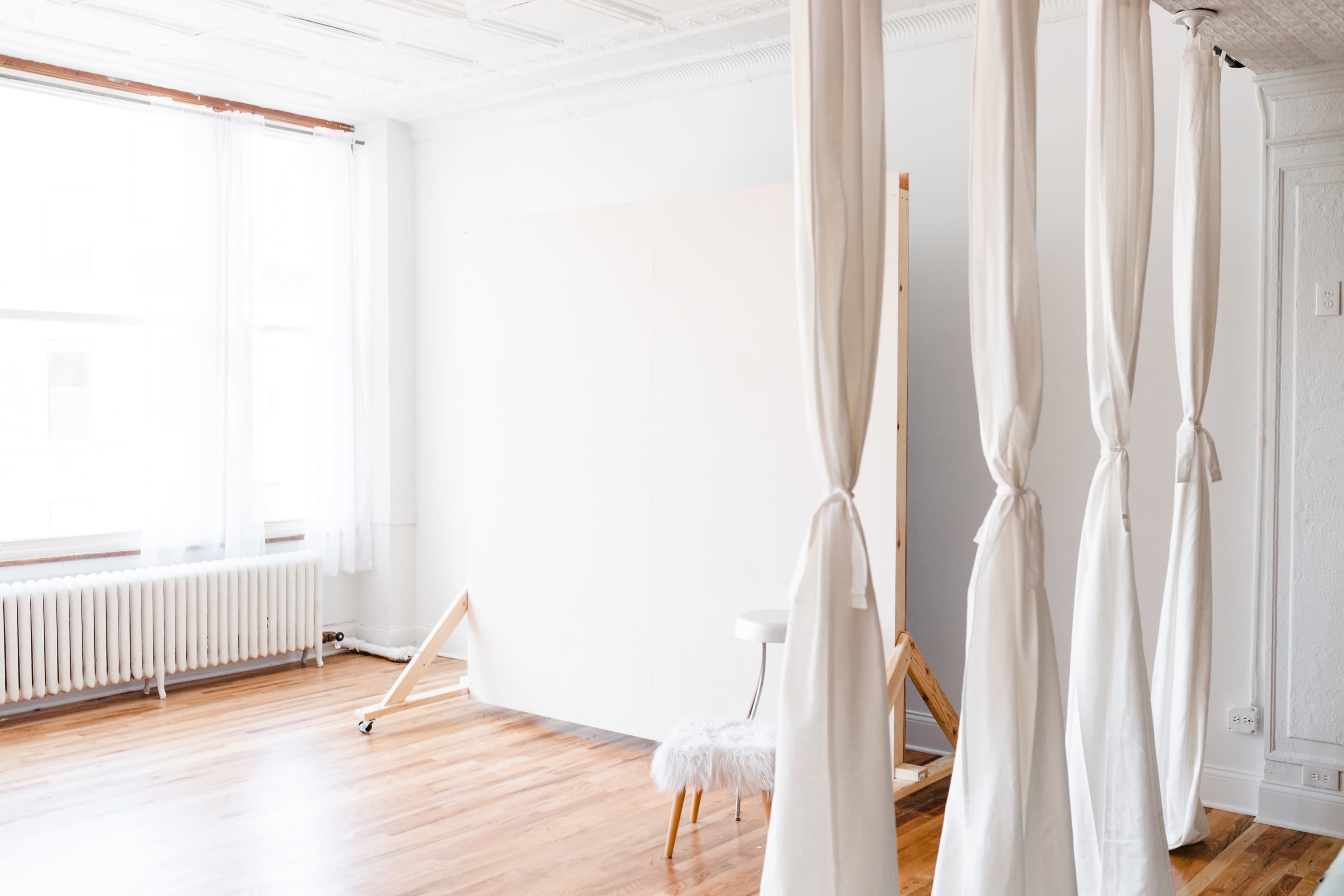 A spacious, well-lit studio with white walls, hardwood floors, a blank canvas on an easel, and fabric curtains tied back.
