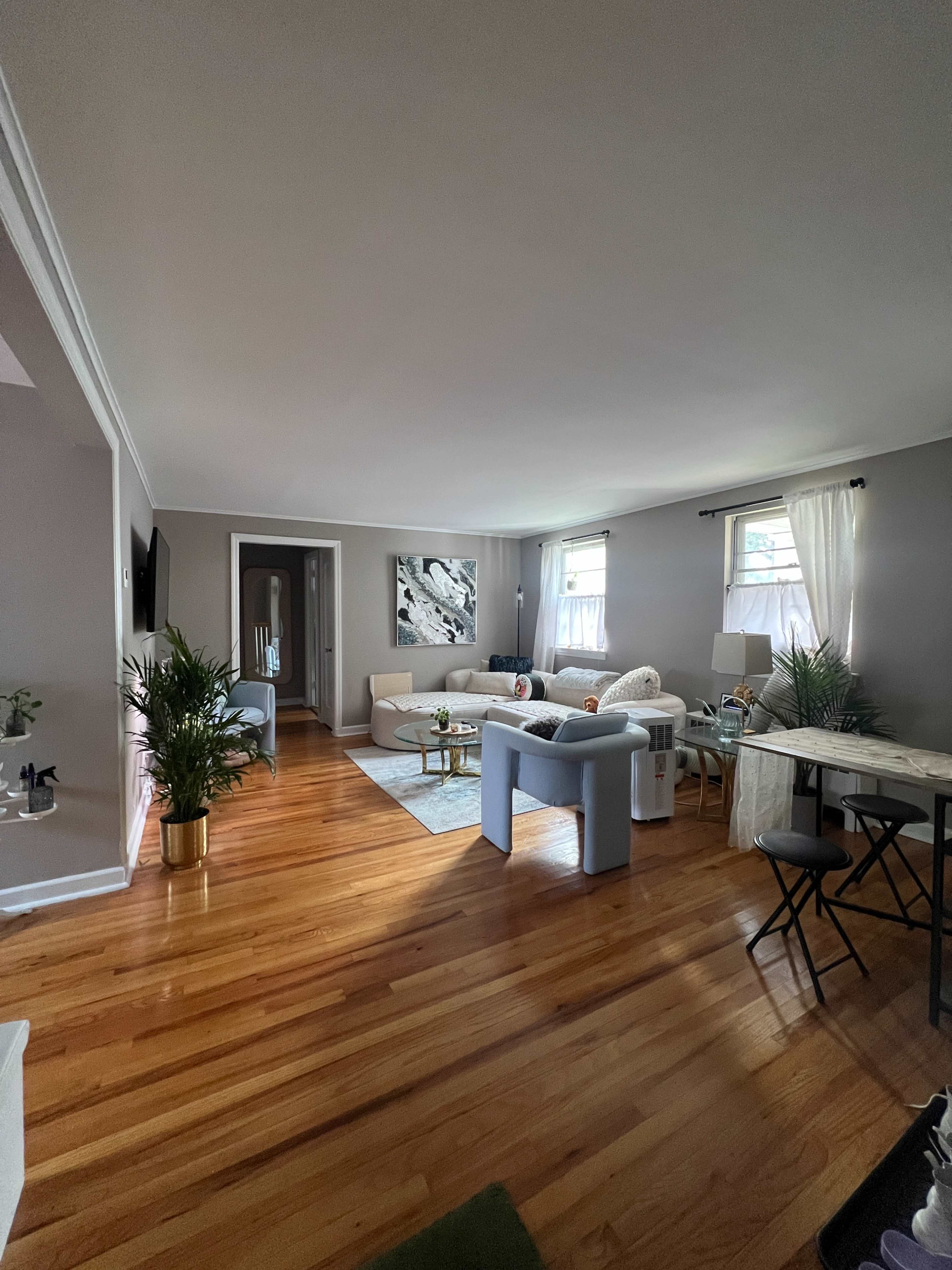 The image shows a well-lit living room featuring hardwood floors, a light-colored sectional sofa, and several plants, with a small dining area visible in the background.