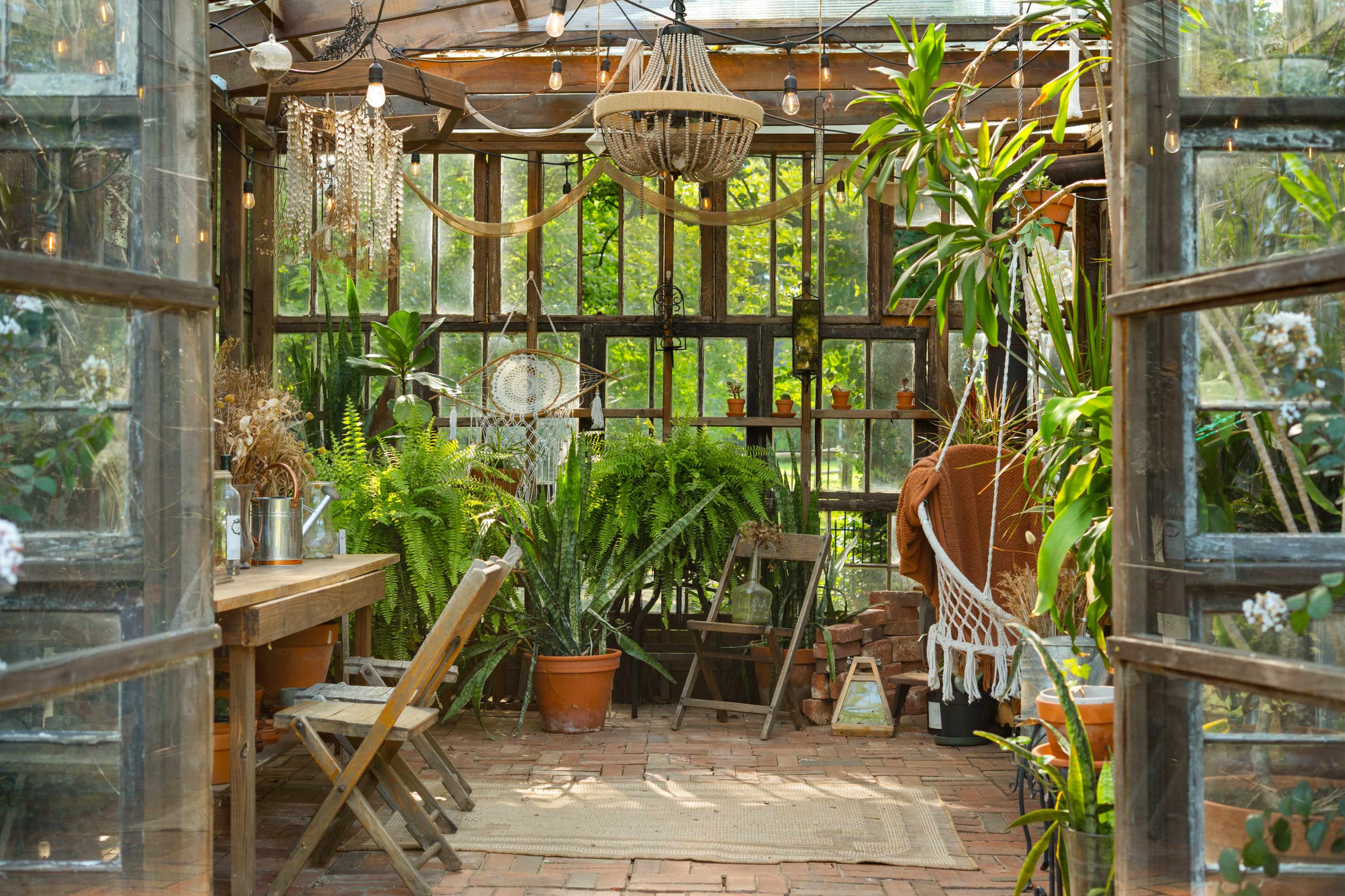 The image shows a cozy greenhouse filled with various plants, wooden furniture, and decorative elements, all illuminated by string lights.