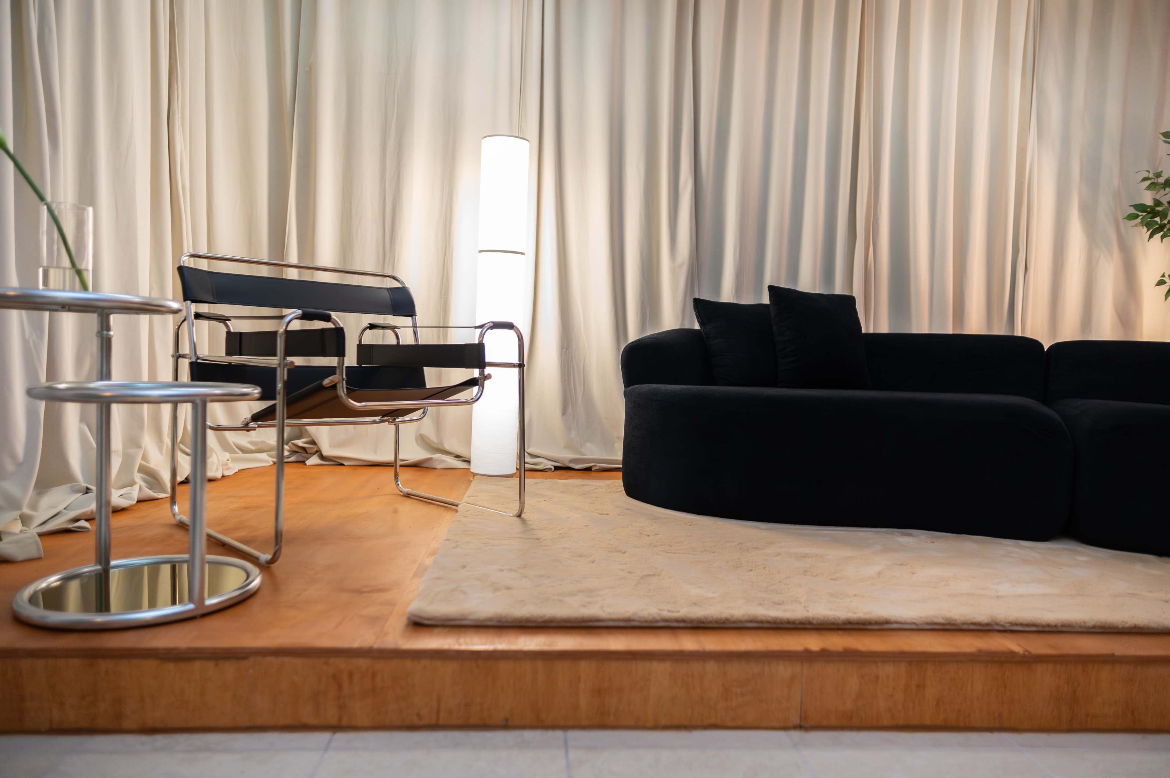 The image shows a modern living space featuring a black sofa, a sleek chair, a floor lamp, and a circular glass table on a light-colored rug, all against a backdrop of soft curtains.
