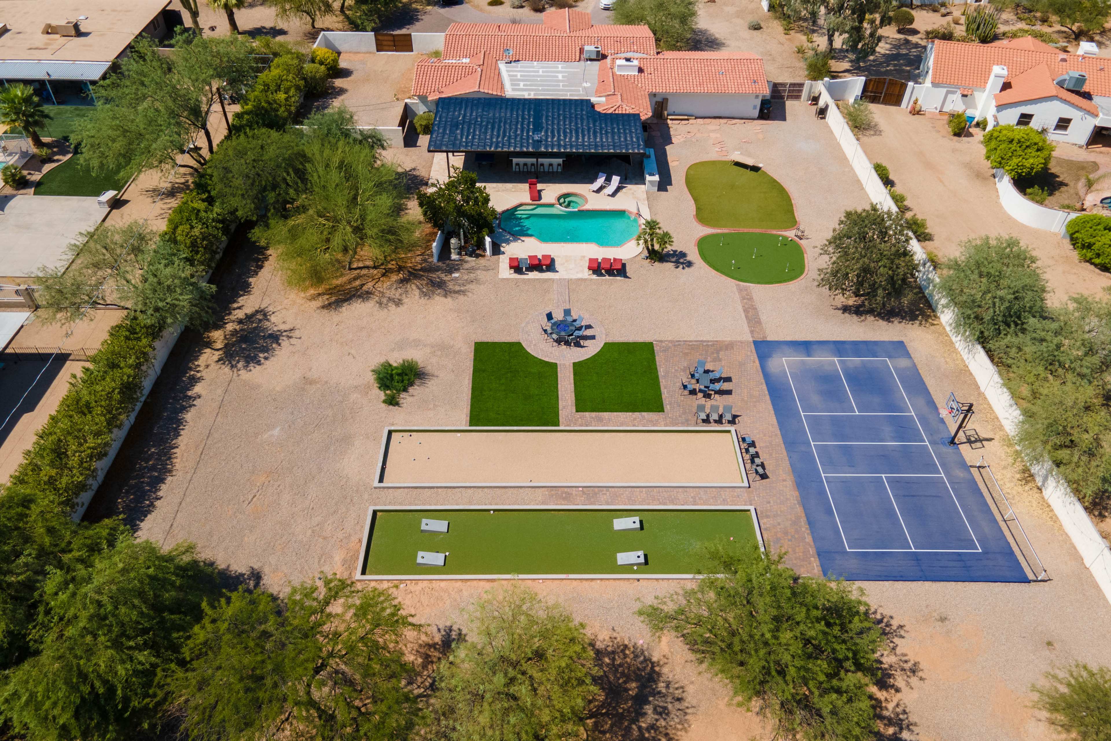The image shows an aerial view of a spacious property featuring a swimming pool, a lawn area, a tennis court, and various seating arrangements.