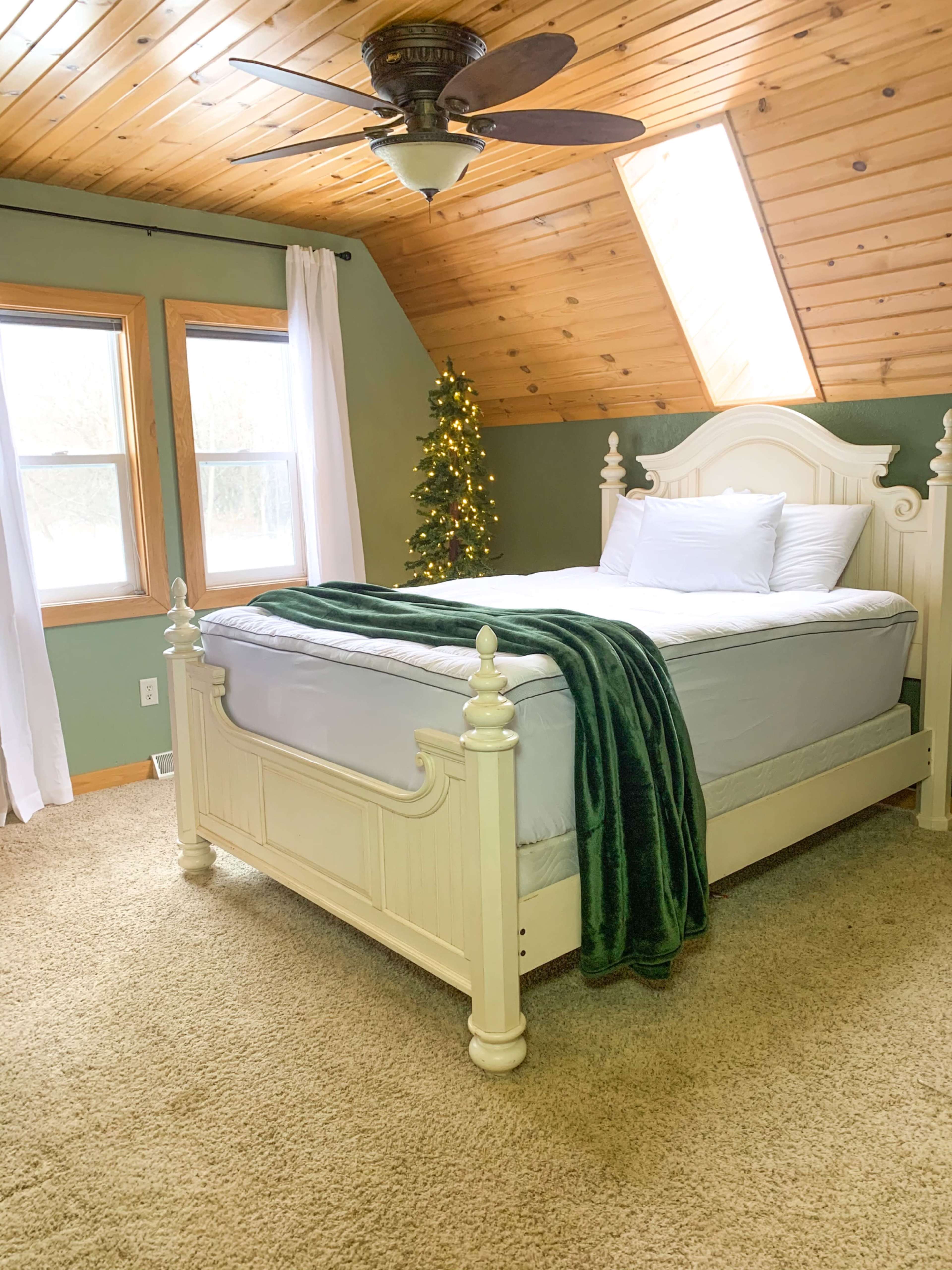 A cozy bedroom features a light-colored wooden bed with a green throw blanket and a small Christmas tree in the corner.