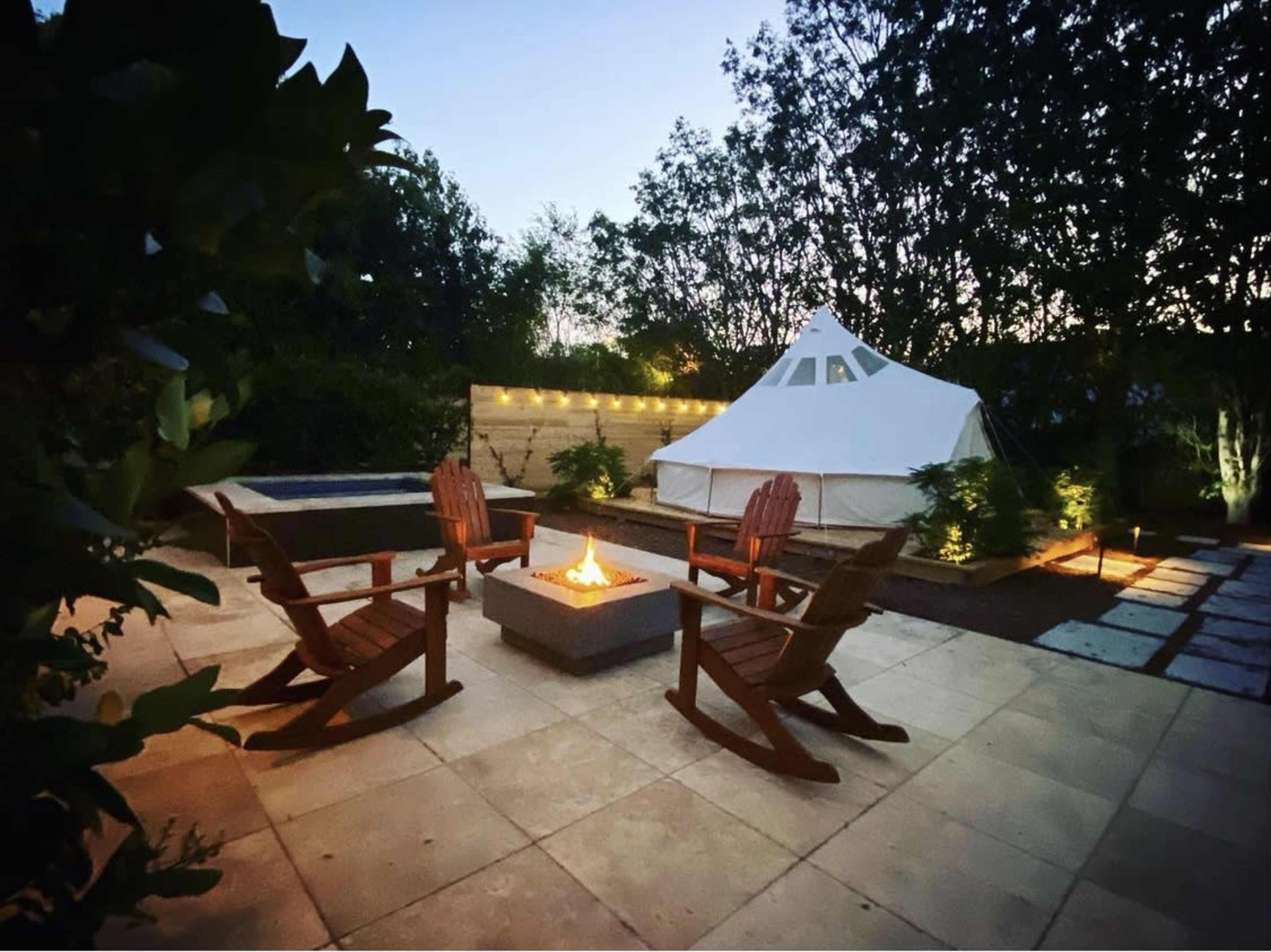 A tent is set up in a backyard with four wooden chairs arranged around a fire pit, surrounded by greenery and stone pathways.
