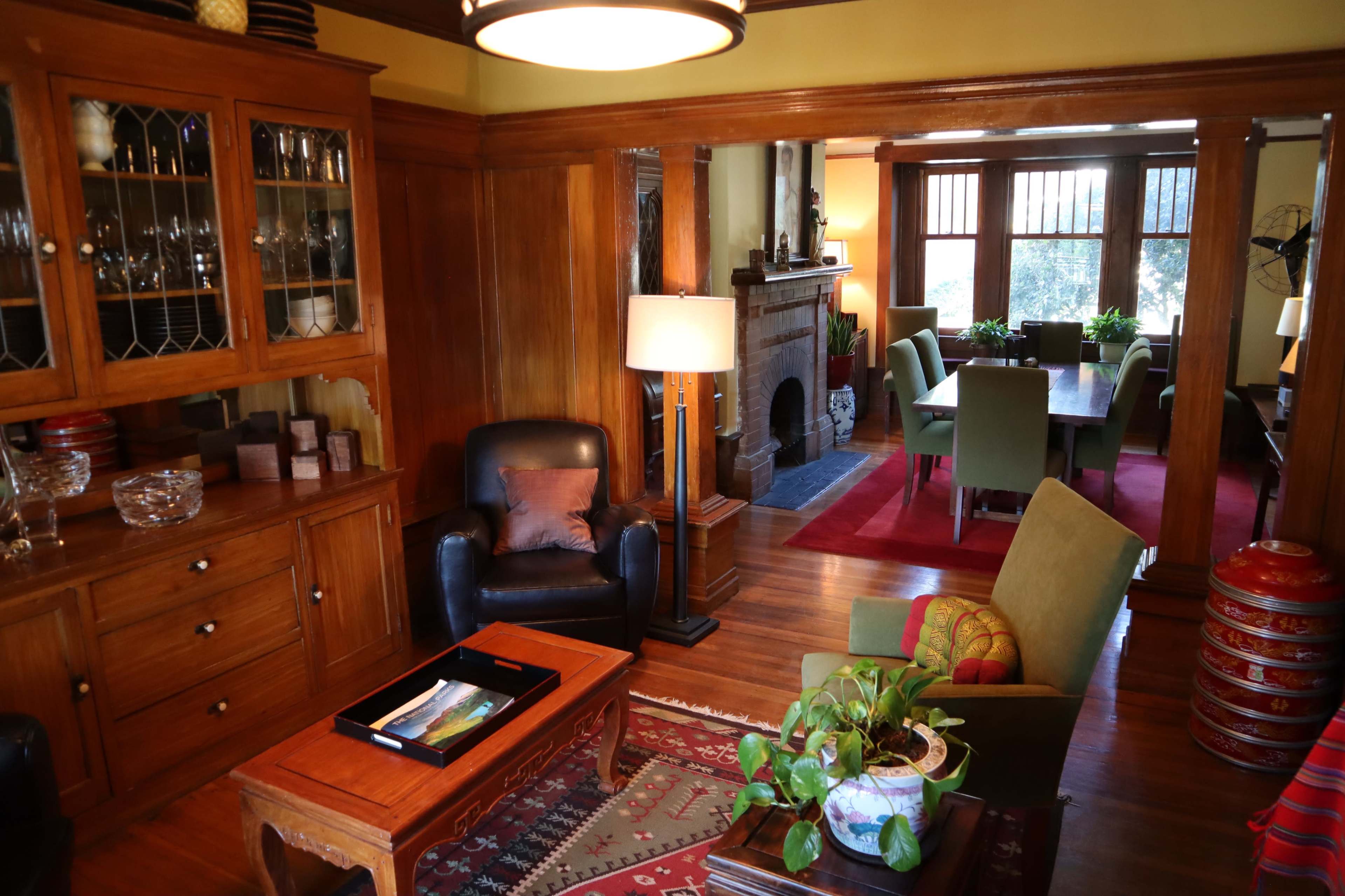 A living room with wooden furniture, a coffee table, a lamp, and a view into a dining area with green chairs and a red rug.