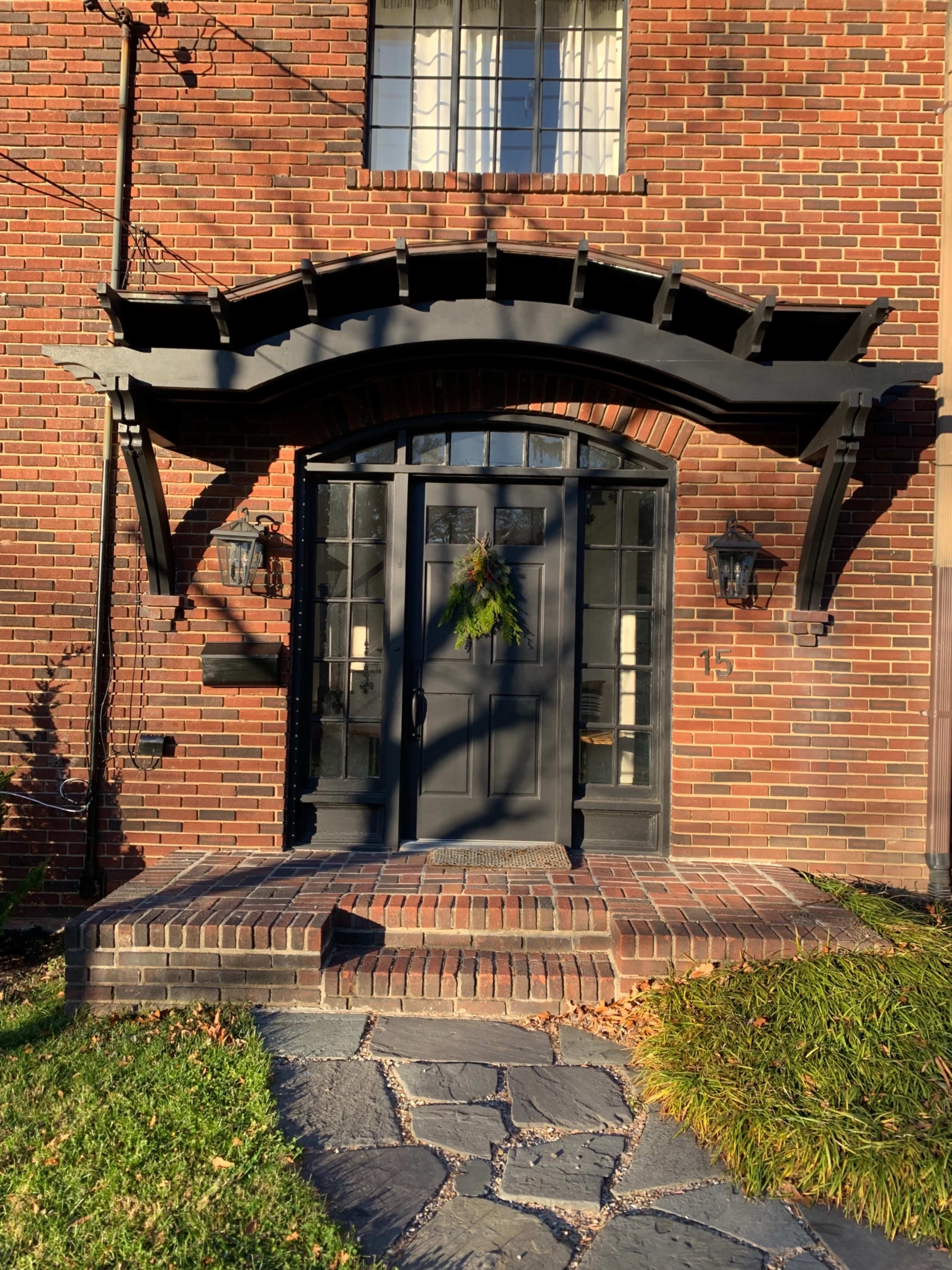 A brick house with a black double door entrance, arched canopy, and a small wreath on the door, surrounded by a stone walkway and green grass.