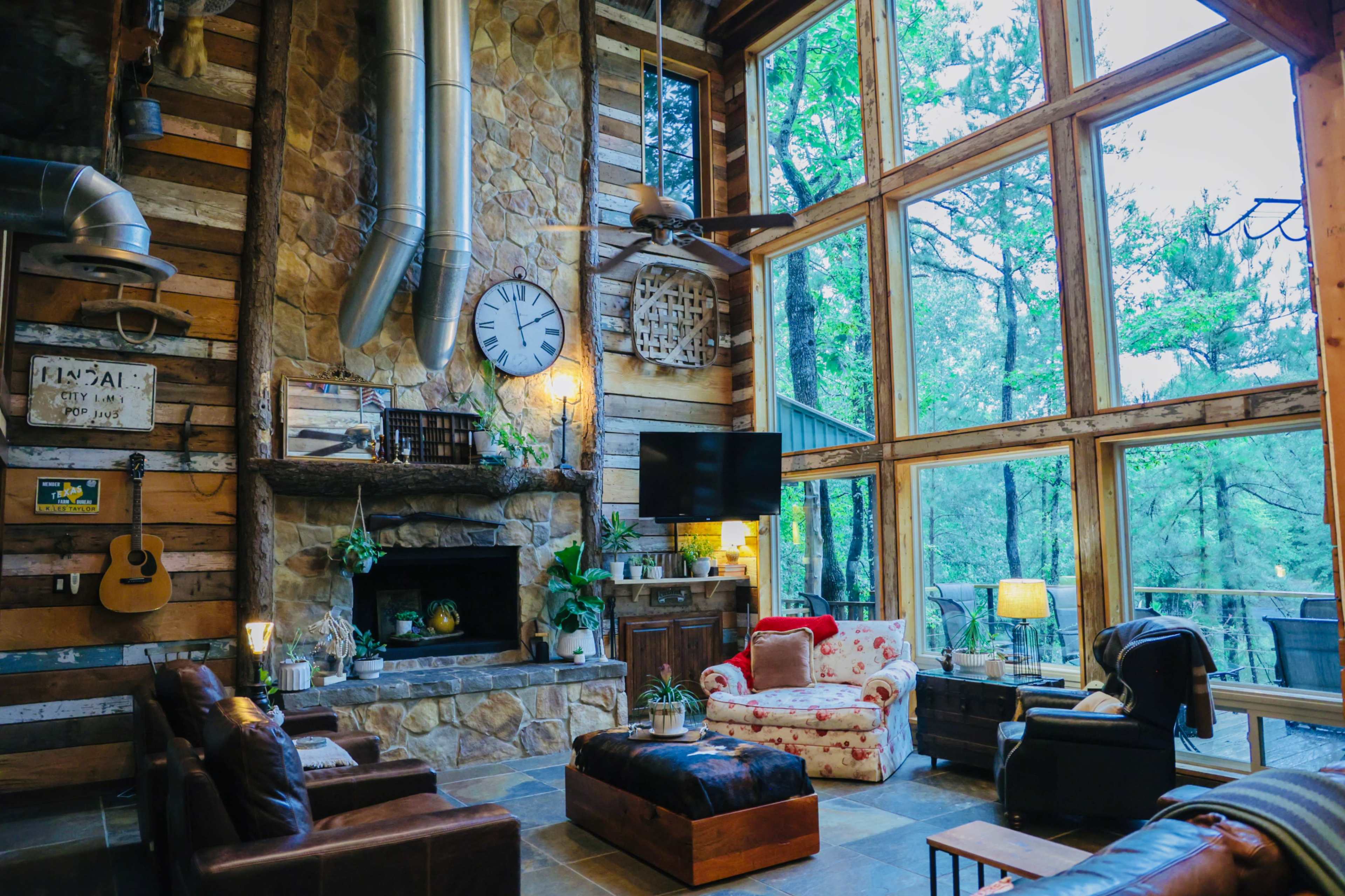 A cozy living room with a stone fireplace, large windows overlooking trees, and rustic wooden walls, featuring a mix of furniture including a floral chair and a dark leather sofa.