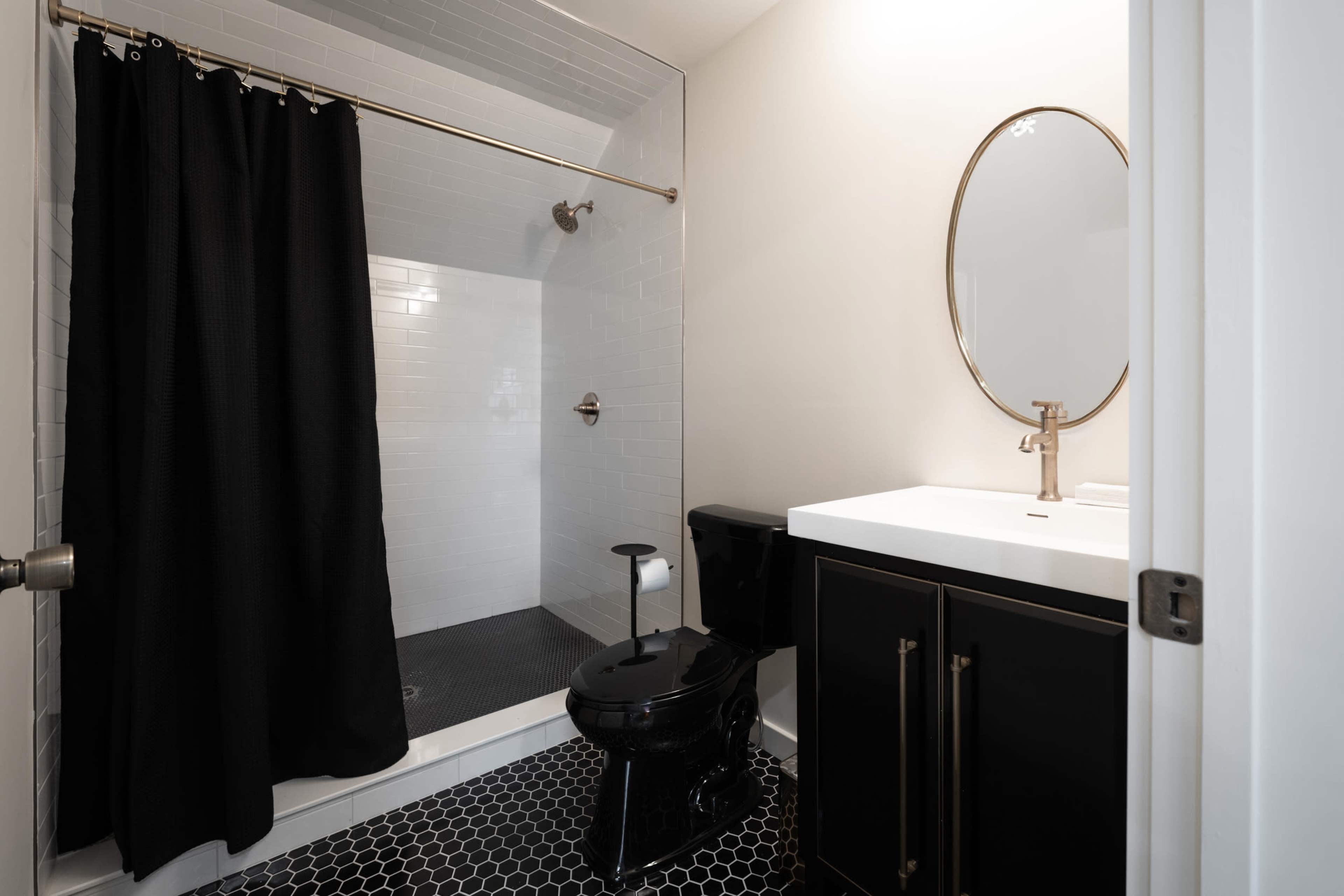 A modern bathroom featuring a black shower curtain, a round mirror above a white sink, and a black toilet, with hexagonal black and white tiles on the floor.