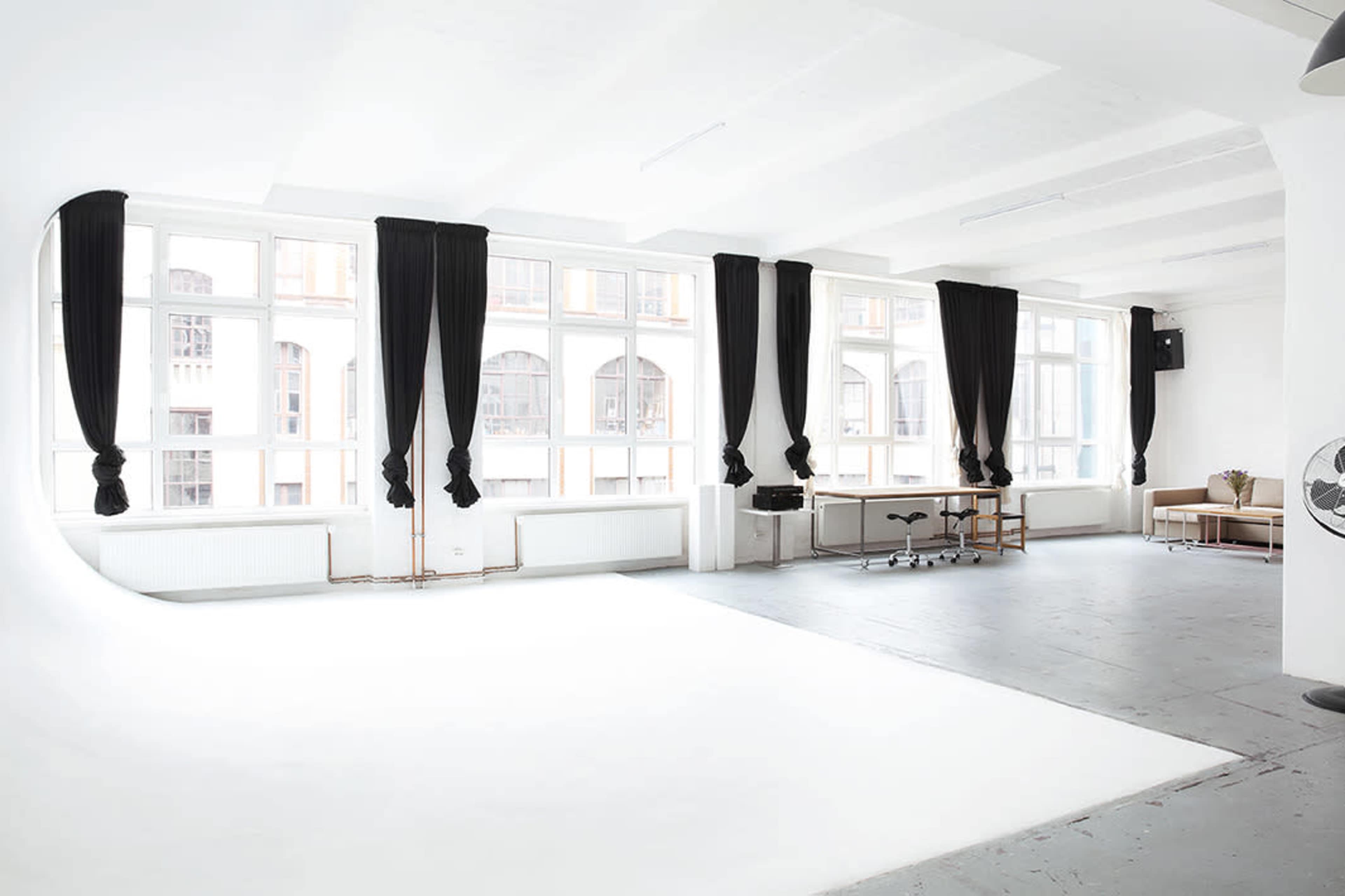 A spacious, well-lit studio with large windows, dark curtains, and minimal furniture including a desk and a fan.