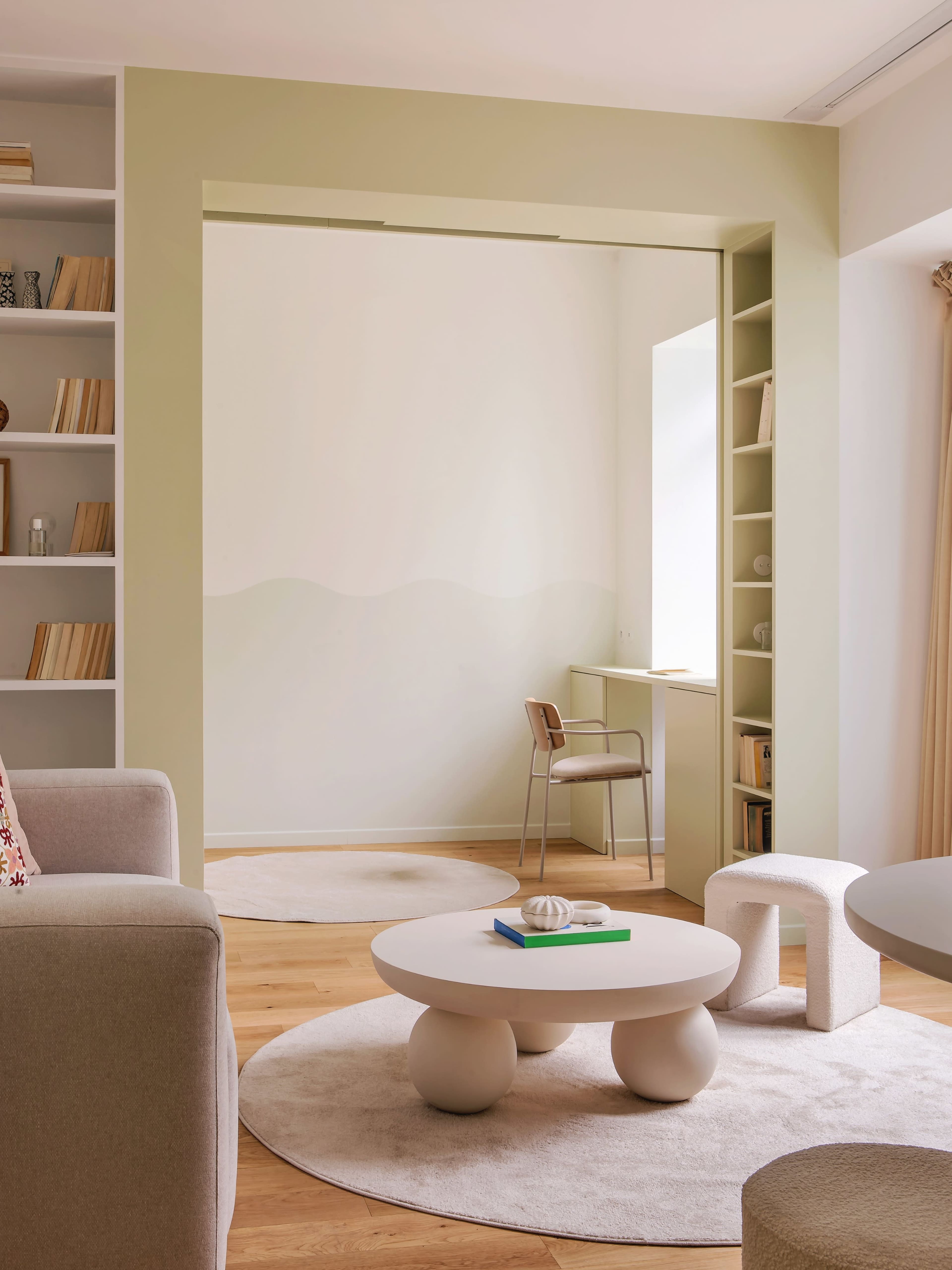 The image shows a modern, minimalistic living space featuring a round coffee table, a small desk, and light-colored rugs on wooden flooring.