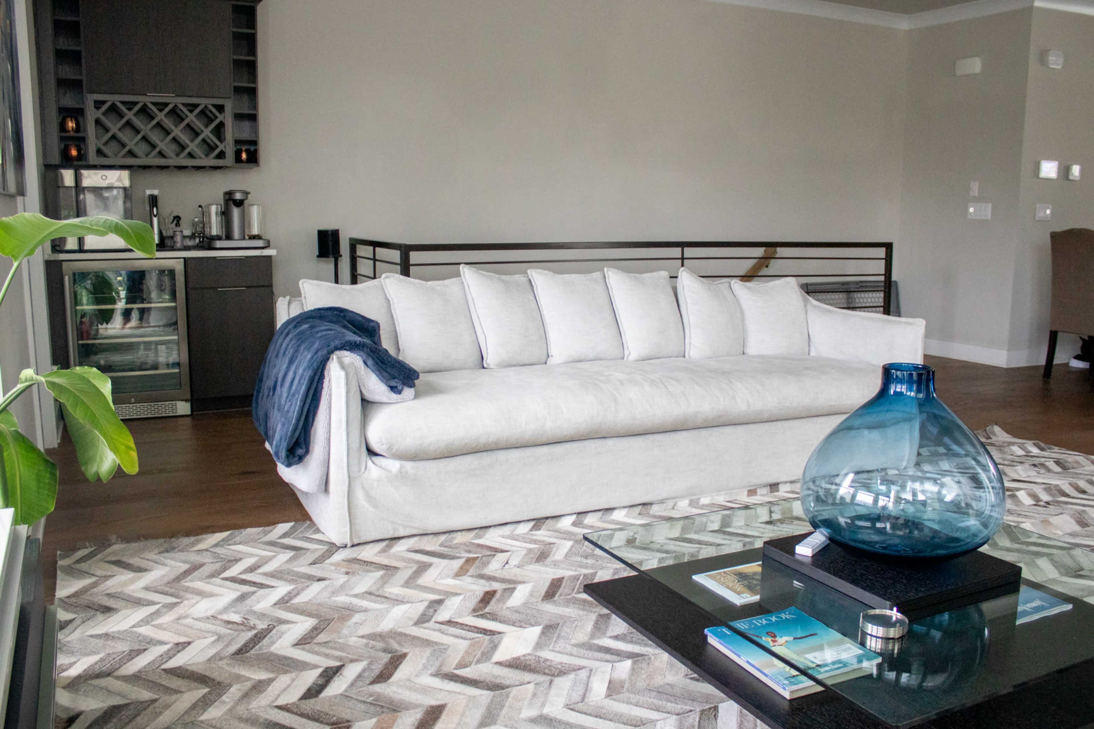 A modern living room featuring a light-colored sofa, a glass coffee table, and a patterned rug, with a kitchen area visible in the background.