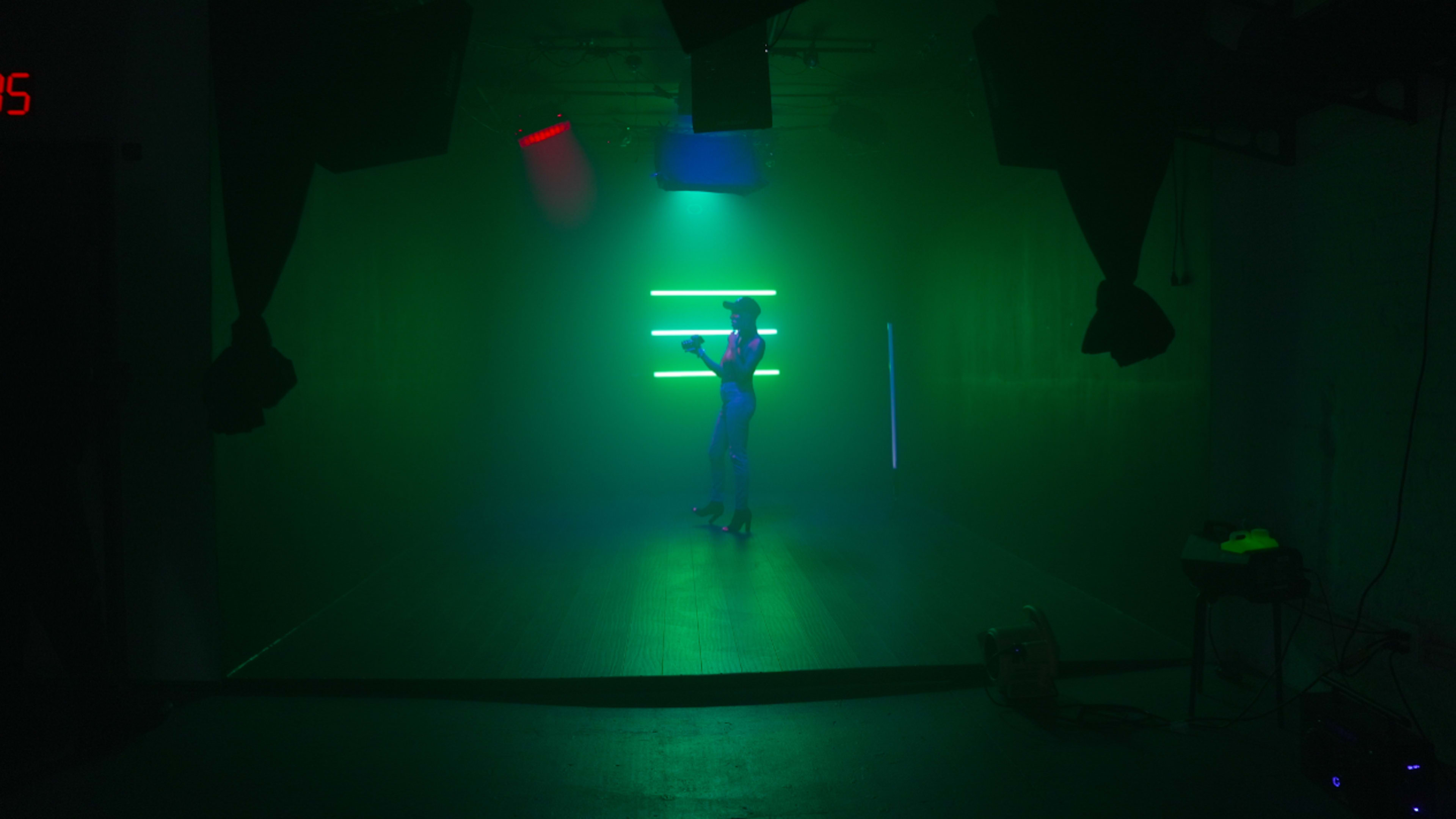 A figure in high heels stands against a backdrop of green and red lights in a darkened studio.