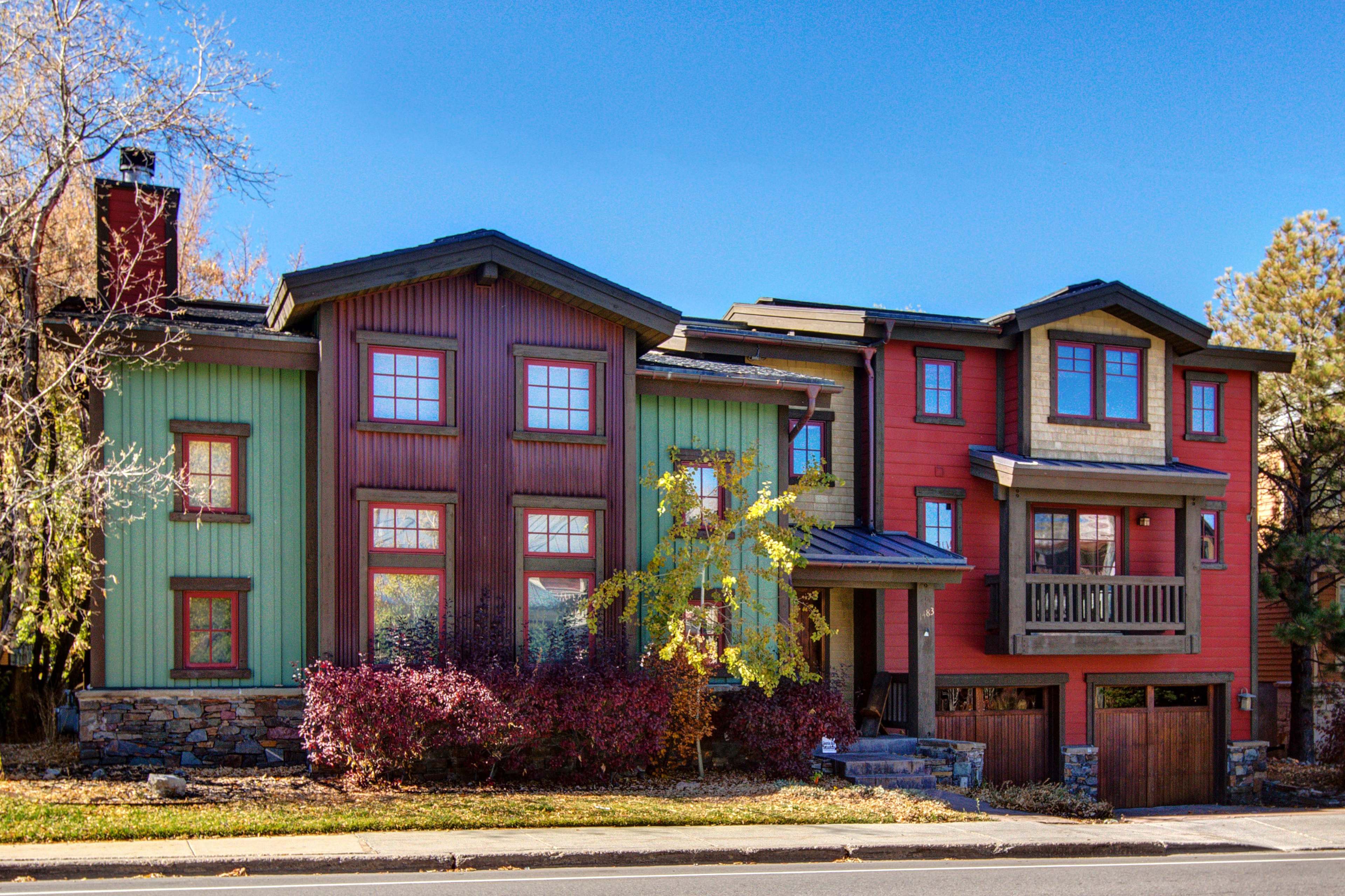 A two-story modern house featuring a combination of green, red, and brown wooden siding with multiple large windows and a front porch.