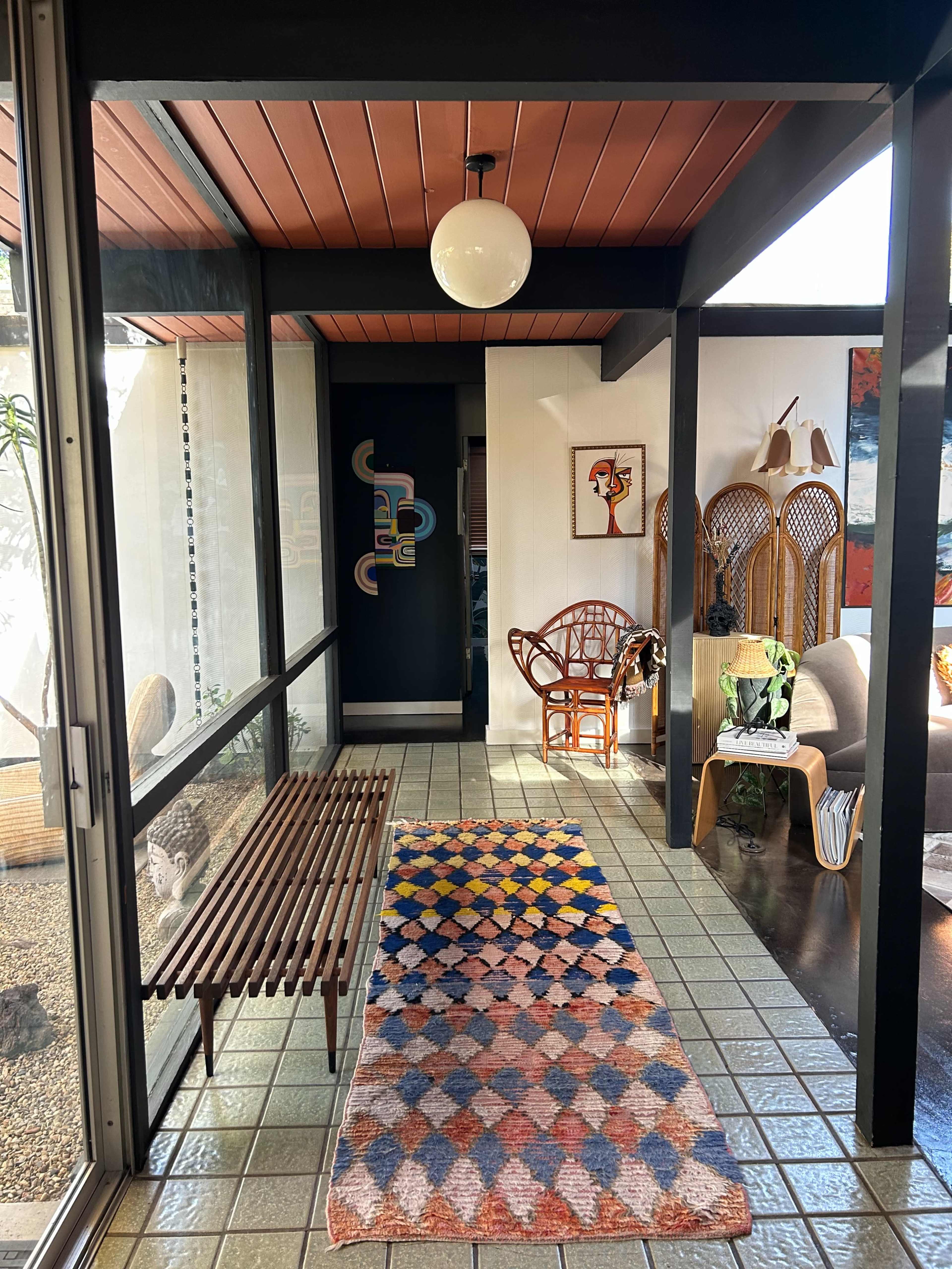 A hallway features a patterned rug, a wooden bench, and a rattan chair, with artwork and plants in the background.