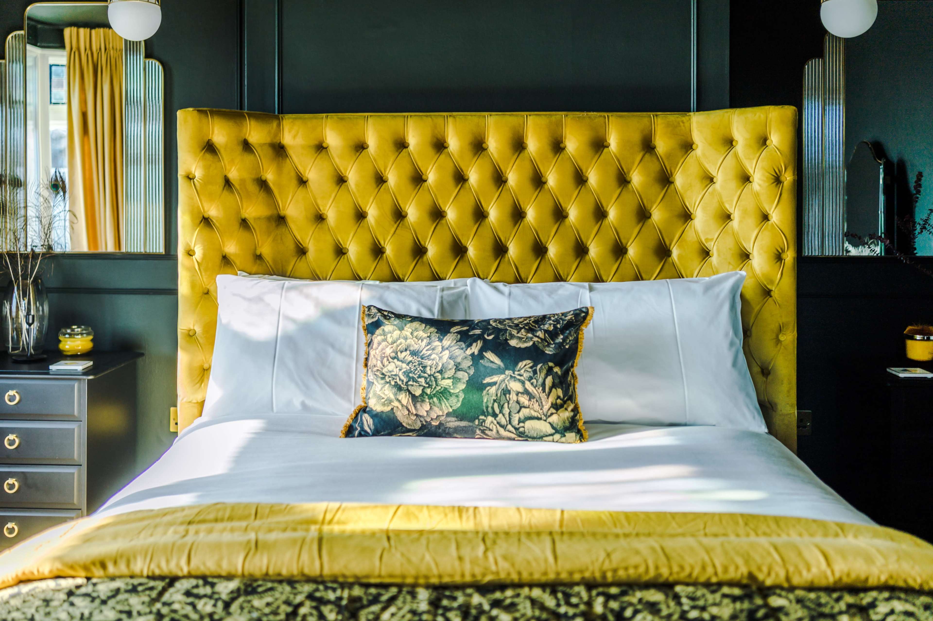 A bedroom with a large yellow tufted headboard, white bedding, and a decorative pillow on the bed, surrounded by dark walls and warm lighting.