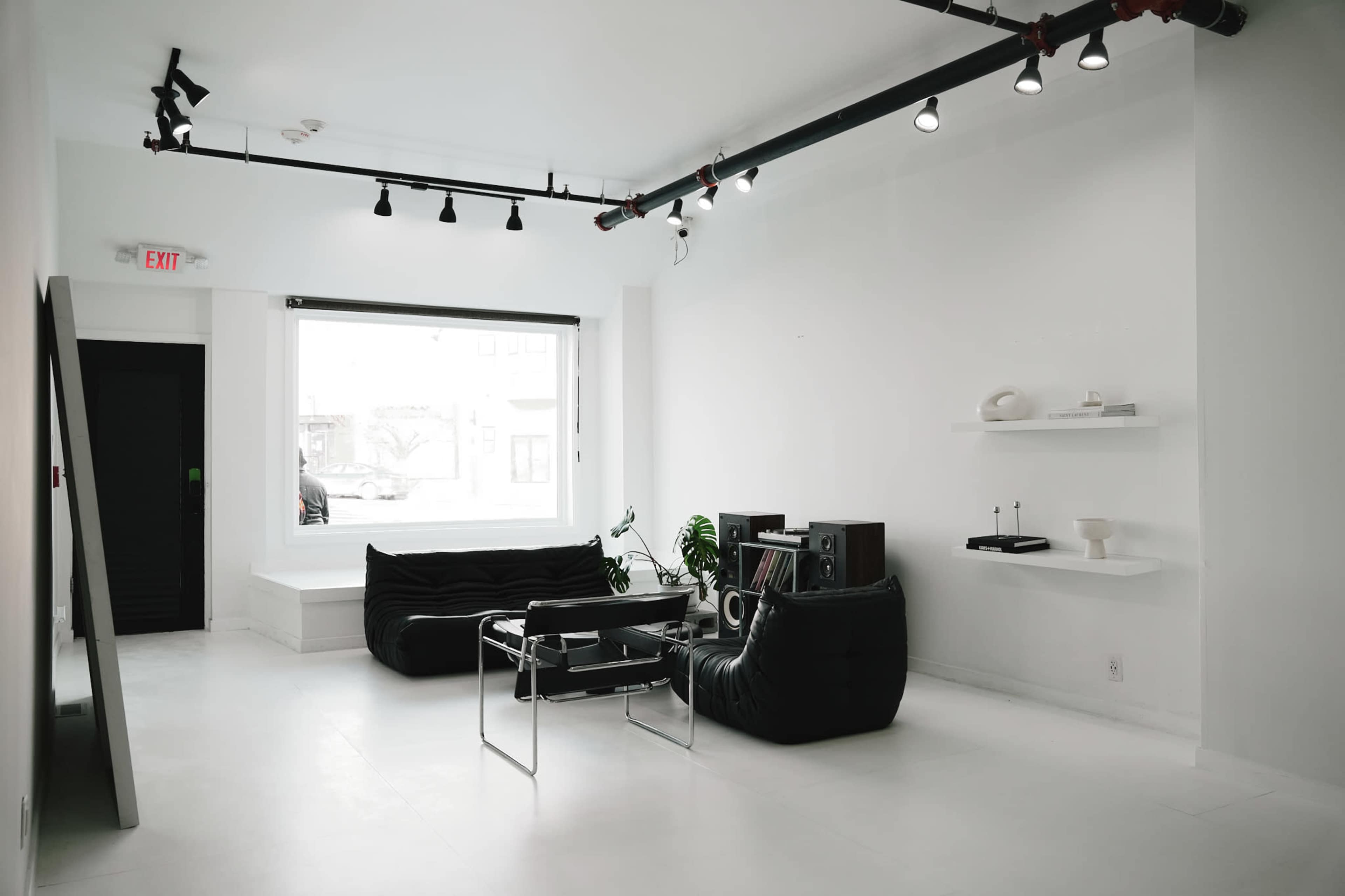 The image shows a minimalist interior of a white-walled room featuring a black couch, a glass coffee table, and a shelf with decorative items.