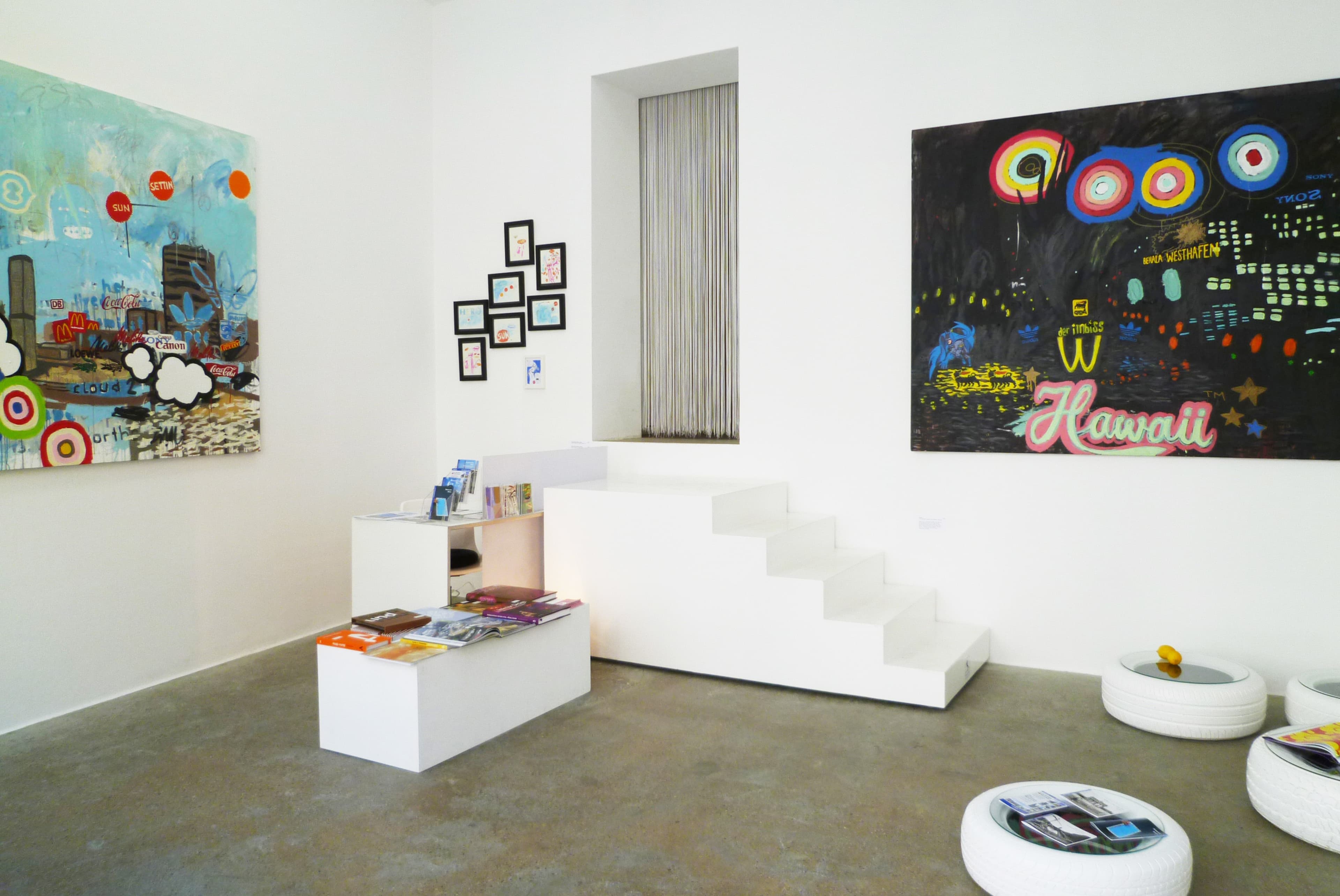 The image shows a brightly lit art gallery featuring colorful paintings on the walls, a set of white stairs leading to a window, and a white table in the center with magazines and brochures.
