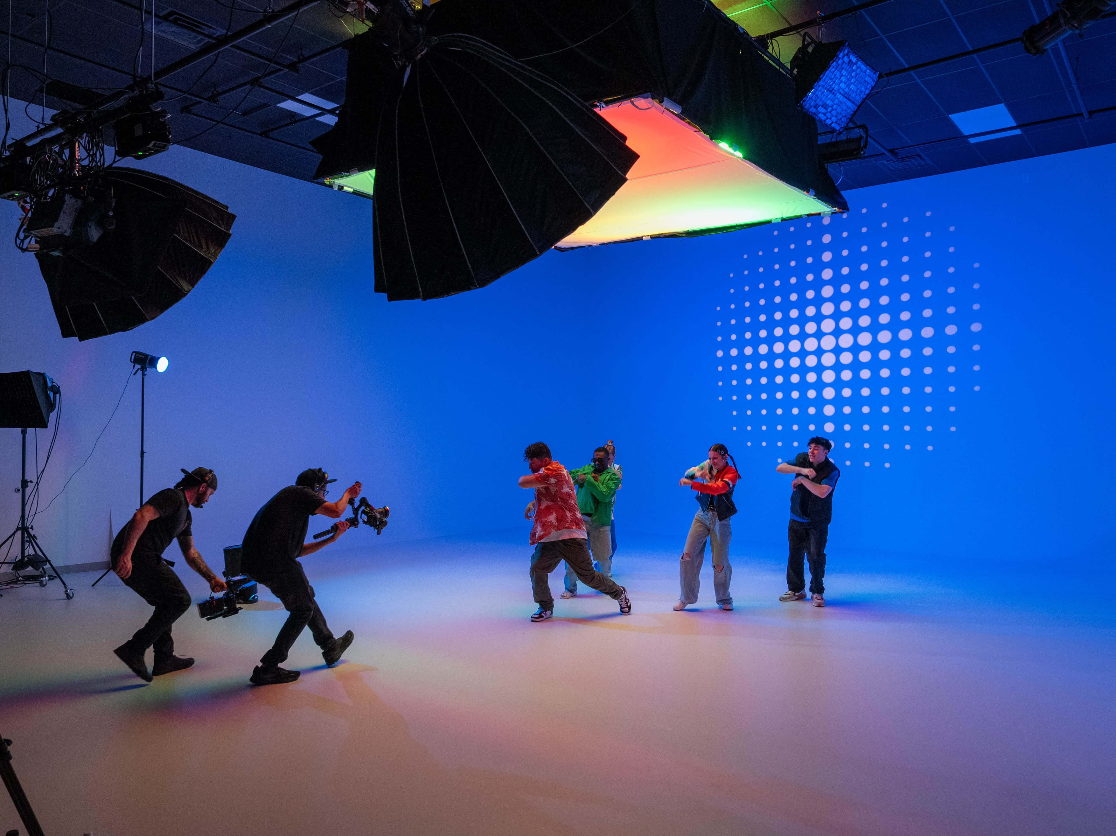 A group of dancers performs in a brightly lit studio with colorful lighting and cameras recording their movements.