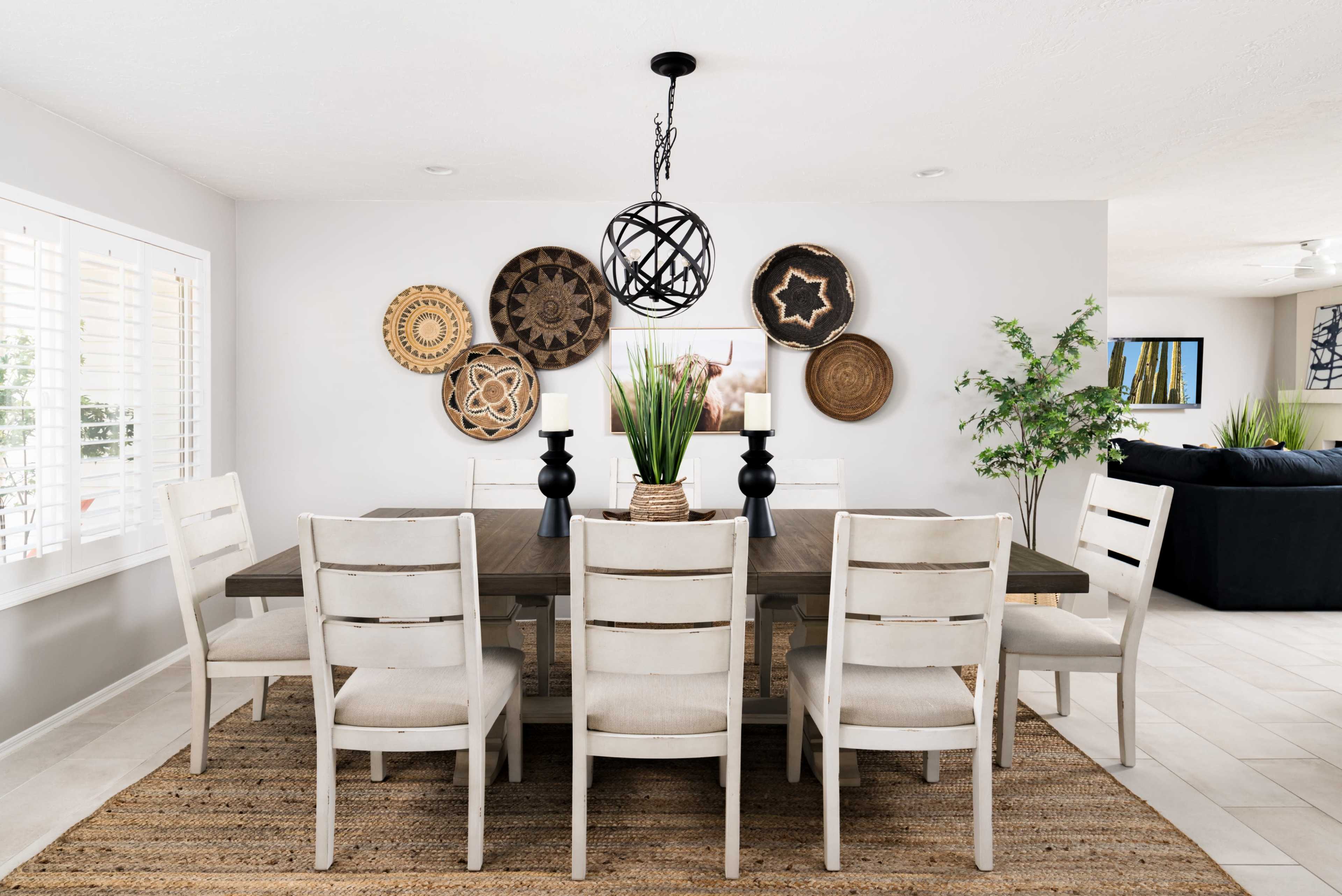 A modern dining room features a large wooden table surrounded by white chairs, with decorative wall hangings and a plant centerpiece.