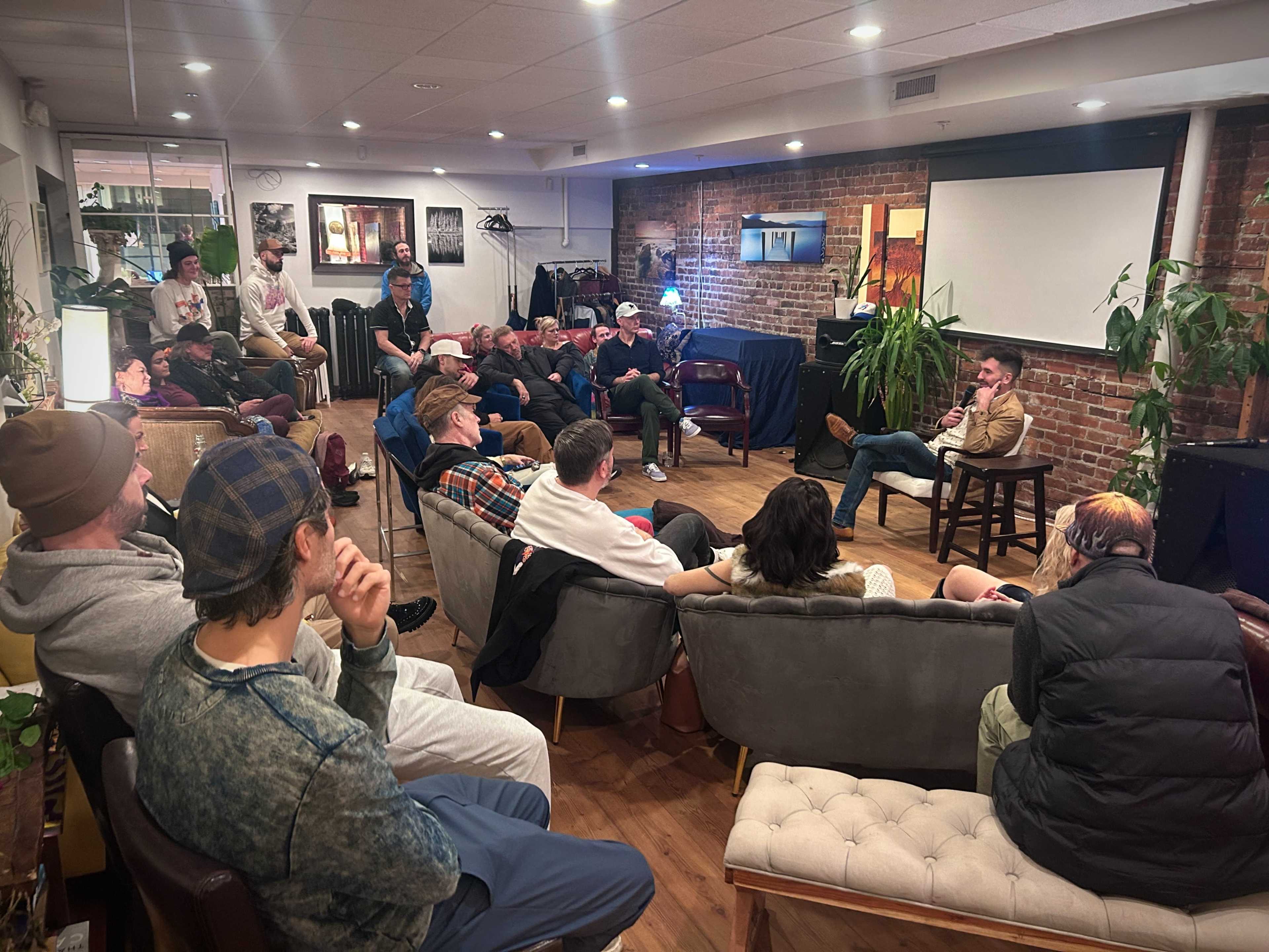 A group of people is seated in a room with exposed brick walls, attending a discussion featuring a speaker on a stage.