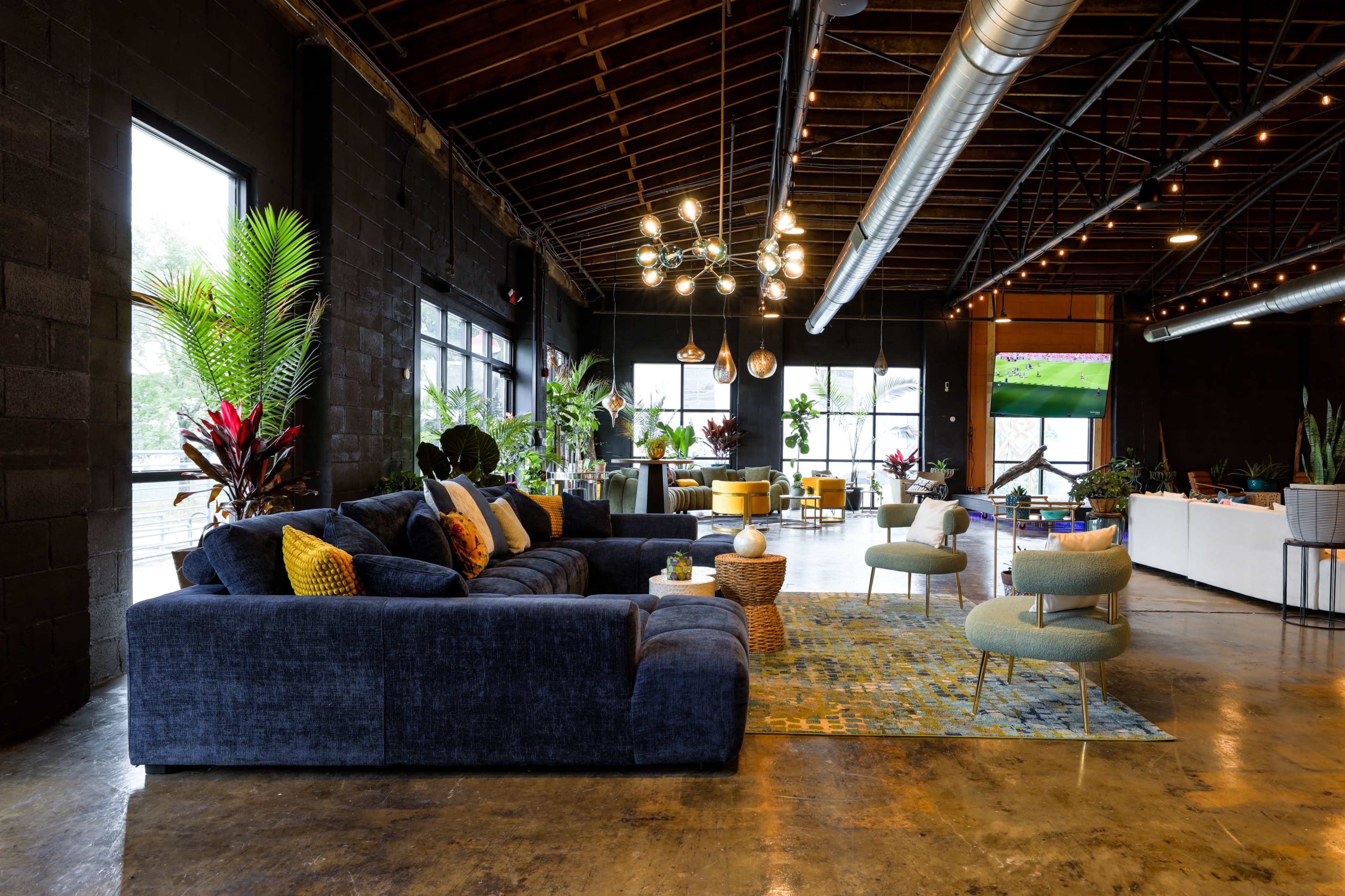The image shows a spacious interior featuring a large blue sectional sofa, stylish chairs, and vibrant plants, with large windows and modern lighting fixtures overhead.